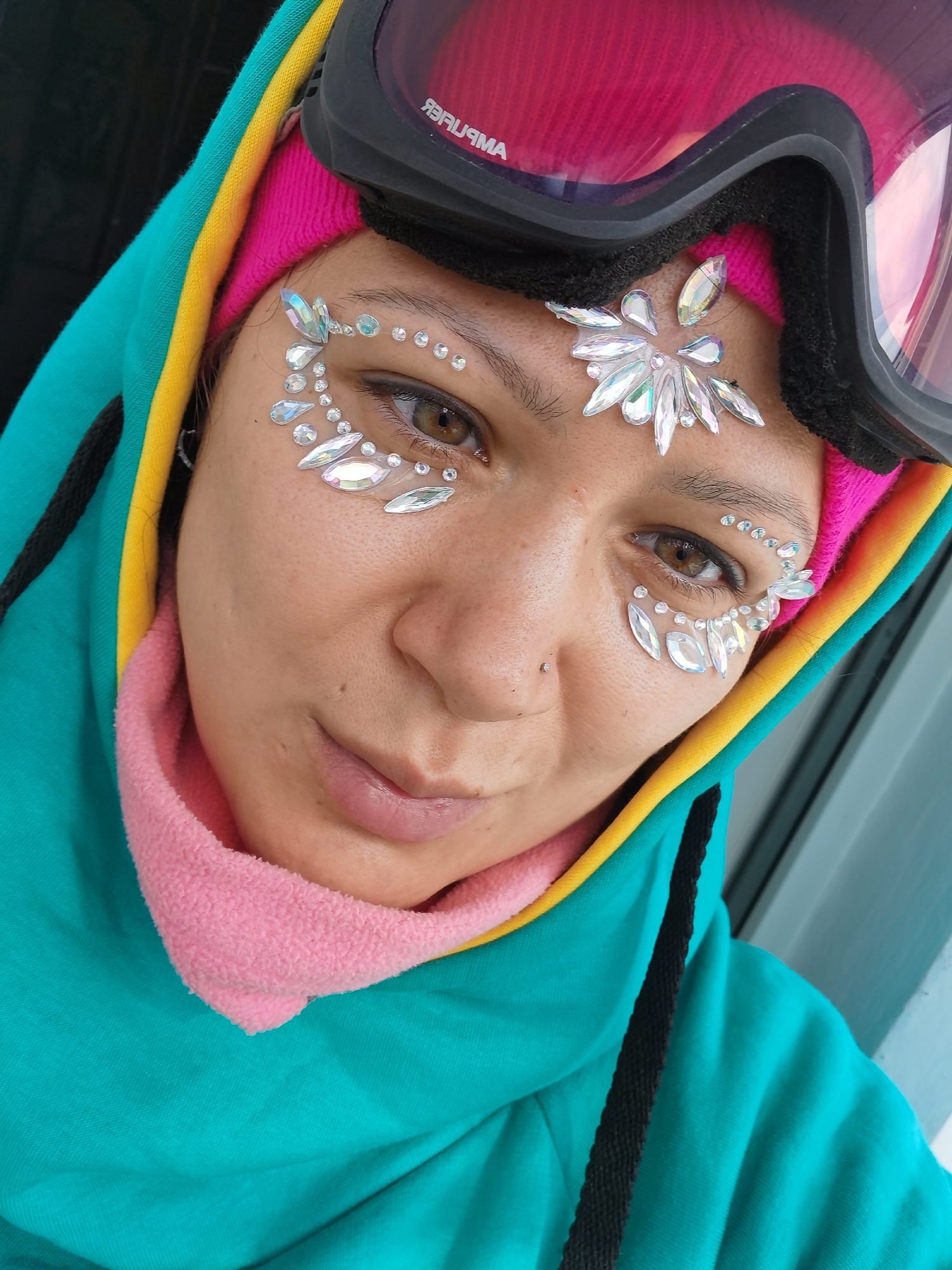 A close-up shot of a fair-skinned individual with brown eyes, adorned with an arrangement of rhinestones around their eyes and forehead, including a larger, flower-like rhinestone between the eyebrows. They are wearing pink goggles. The individual is wearing a multi-layered, hooded sweatshirt with colors including blue, yellow, and pink. A matching pink fleece collar is visible. A nose piercing is also visible. The location appears to be in an outdoor setting, in the city of Unknown, Unknown.