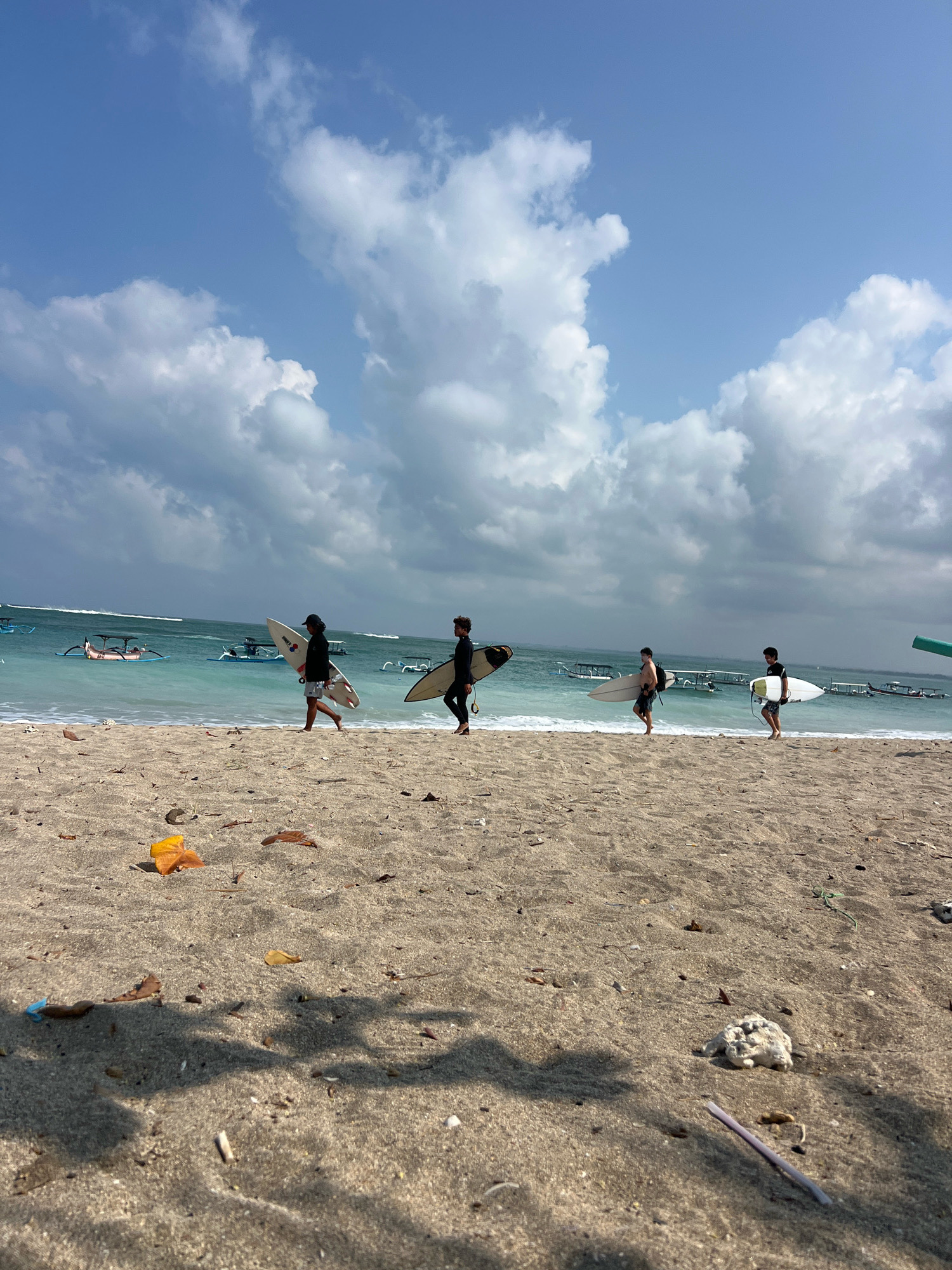 In Kuta, Indonesia, a group of four surfers are walking along a sandy beach, each carrying a surfboard. The surfers are spaced apart and walking in the same direction, away from the water. The ocean is calm with small waves breaking near the shore. Several traditional Balinese fishing boats are visible in the turquoise water. The sky is partly cloudy with fluffy white clouds against a bright blue backdrop. The sand in the foreground is coarse and littered with small pieces of debris, including leaves and plastic. The overall scene depicts a leisurely afternoon at the beach, with the surfers heading to or from a surfing session.