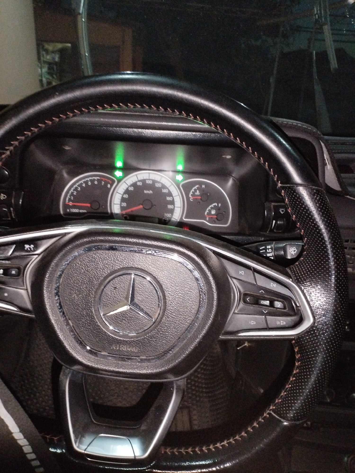 The image shows the interior of a car, focusing on the steering wheel and dashboard. The steering wheel is black with brown stitching and features a Mercedes-Benz logo in the center. There are various buttons on the steering wheel, suggesting it is equipped with controls for audio, cruise control, or other vehicle functions. The dashboard displays two main gauges: a tachometer on the left and a speedometer on the right, with smaller fuel and temperature gauges. Several green indicator lights are illuminated on the dashboard. A windshield wiper control stalk is visible to the right of the steering wheel. The overall scene suggests the car is parked or stationary. The image is taken from the driver's perspective. No individuals are visible in the image. The location in Termez, Uzbekistan is not evident from the visual information provided in the image.