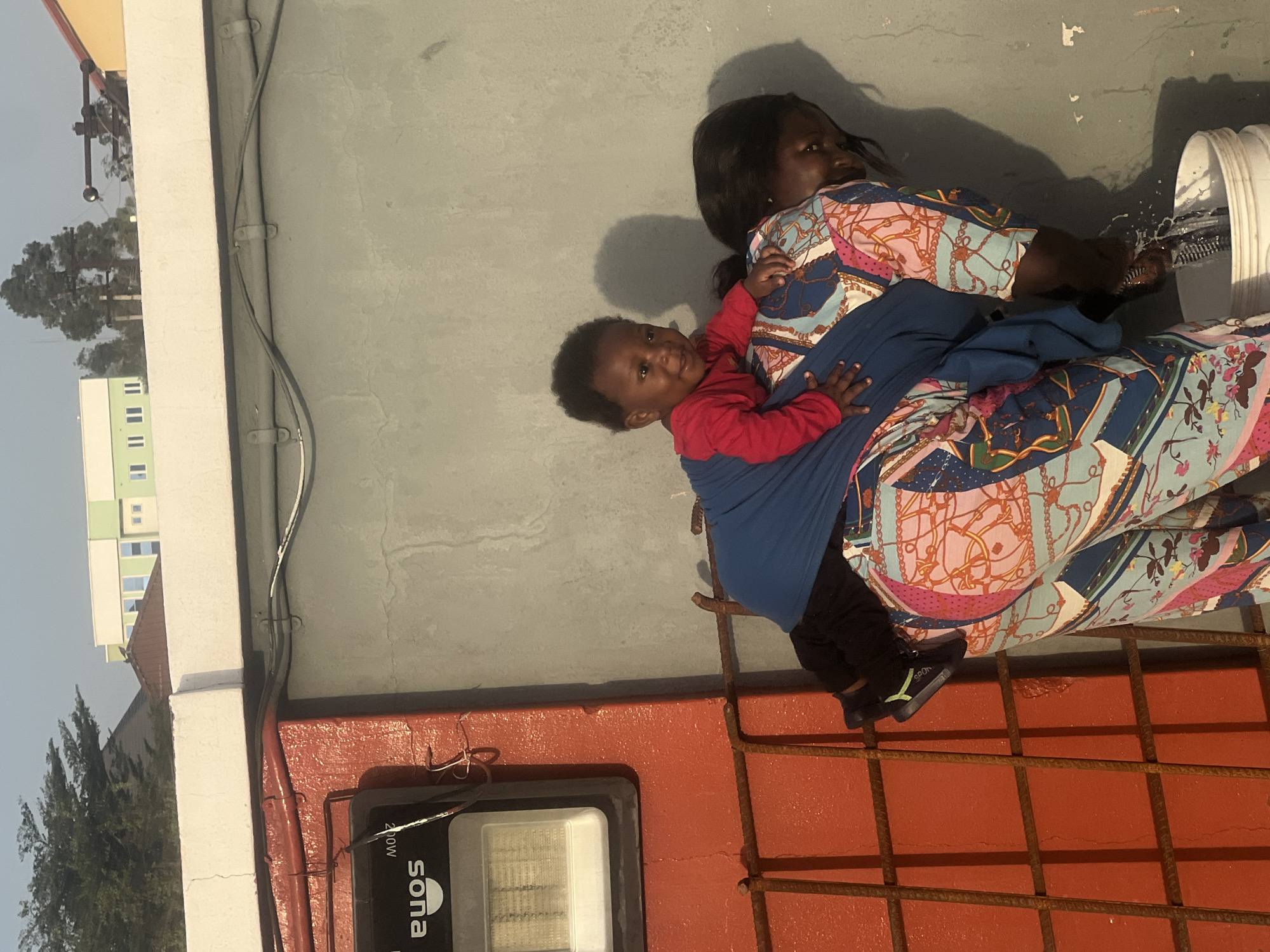 An outdoor scene in Gura Top, Nigeria, captured in bright daylight, shows a woman carrying a young child on her back while engaged in a domestic task. The woman, dressed in a colorful patterned dress with blue, pink, white, and orange chain and floral designs, also has a plain blue fabric wrapped around her. She is seen from the waist up, leaning over a white bucket from which water is splashing, indicating she is washing something. Her right hand is partially visible, immersed in the bucket.
The young child, positioned on the woman's back and secured by the fabric, wears a red long-sleeved shirt and black trousers with black and green shoes. The child looks directly at the camera with a pleasant, smiling expression.
The setting appears to be an elevated concrete area, possibly a rooftop or balcony, with a grey wall behind the subjects. In the background, under a clear sky, are green trees and distant multi-story buildings with visible windows. A red-painted surface, possibly a parapet or ledge, is visible in the foreground, featuring a metal grate made of rebar. A light fixture is mounted on this red surface, displaying the text "sona" and "210W". The word "SPON" is visible on the child's shoe. Electrical wiring is secured along the grey wall. The presence of strong shadows suggests bright, direct sunlight.