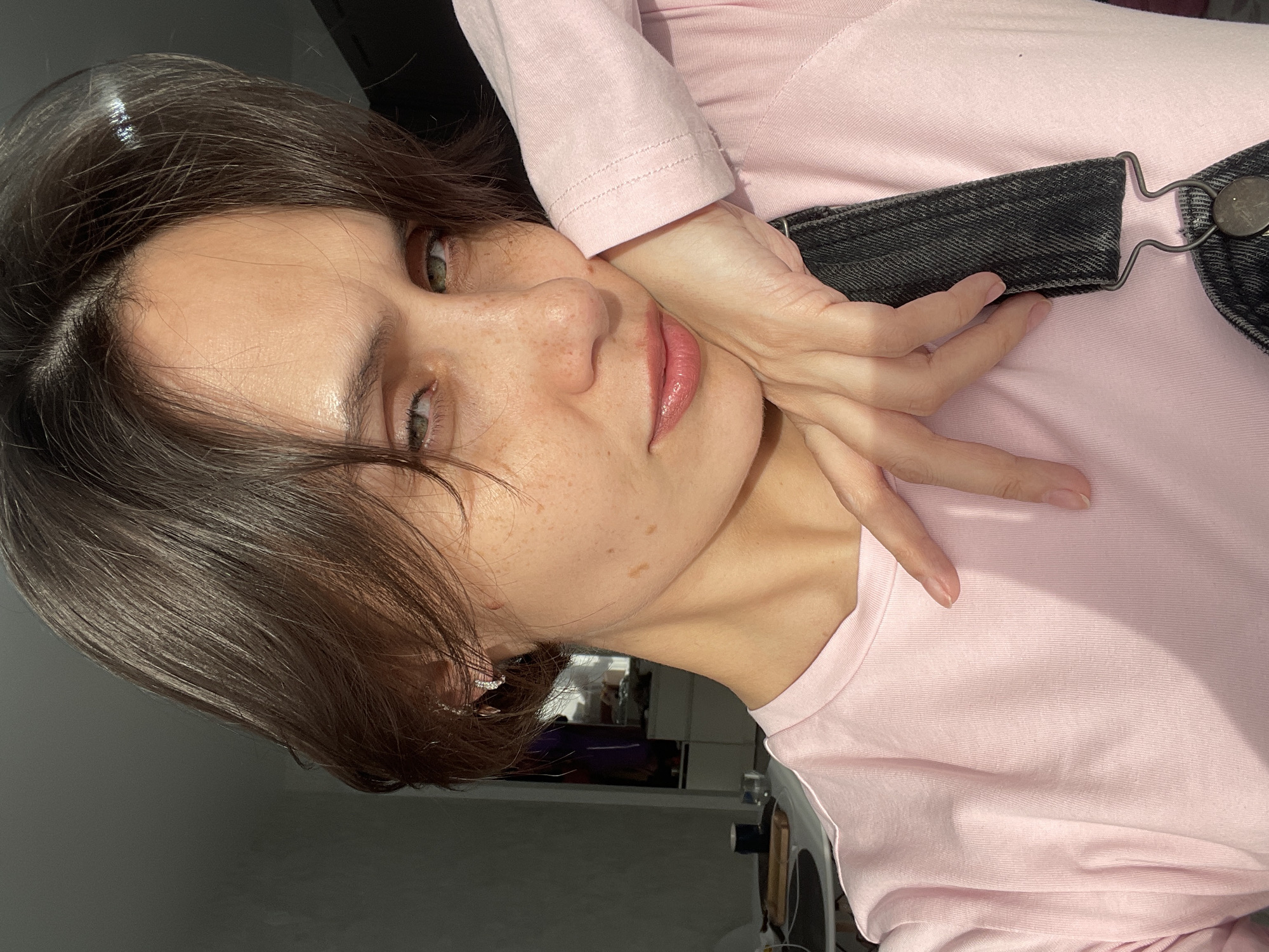 A medium close-up, tilted portrait shows a young woman, likely in Izhevsk, Russia, looking directly at the camera. She has dark brown, medium-length hair, light-colored eyes (appearing green or hazel), and visible freckles on her nose and cheeks. Her lips are slightly parted and enhanced with a glossy pink hue. She is wearing a light pink long-sleeved top and dark denim overalls, with a strap and metallic buckle visible over her shoulder. Her left hand is gently placed on her cheek, fingers slightly splayed, contributing to a posed and serene expression that carries a subtle allure. Natural daylight illuminates her face and hair, creating highlights and shadows consistent with an indoor setting during the daytime. The background is softly blurred, showing vague outlines of a room. No text is visible in the image.