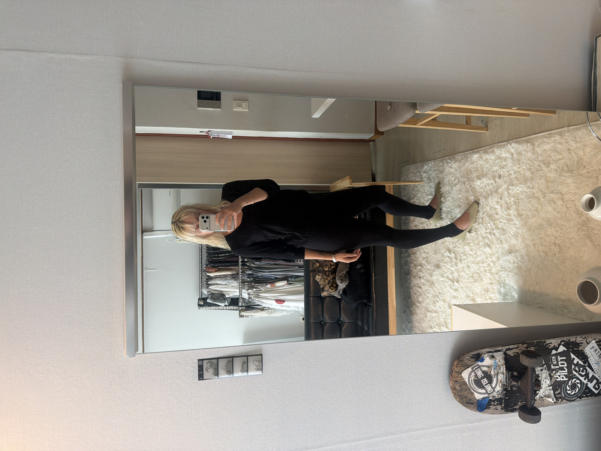 The image is a mirror selfie taken in what appears to be a bedroom or living space in Seoul, South Korea. The reflection shows a woman holding a smartphone, presumably taking the photo. She is wearing a black long-sleeved top and black leggings.
The background reveals details of the room. A wardrobe or clothing rack filled with various clothes is visible. A fluffy white rug covers part of the floor. A chair with wooden legs can also be seen in the reflection. A skateboard is mounted on the wall below the mirror.
The scene is well-lit and appears to be in a modernly decorated apartment or house. The photograph provides no indication of the specific location, but can be attributed to Seoul, South Korea given the location information.
