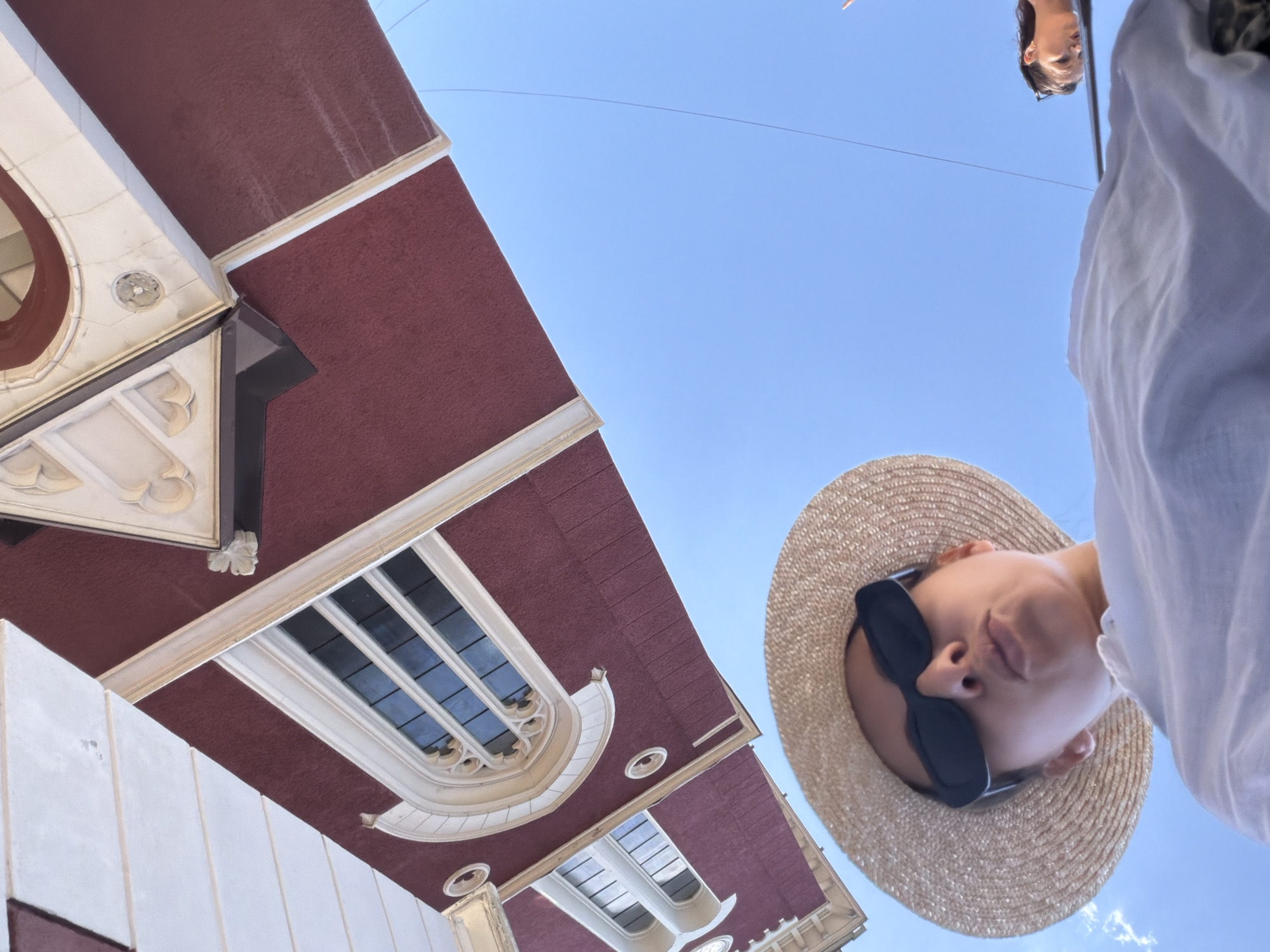 A low-angle, wide shot captures a woman wearing a straw hat and sunglasses, looking directly at the camera with her lips pursed. She is dressed in a white top. Behind her, a portion of a red building with white architectural details and windows is visible, tilted and appearing to hang from the sky. The building's design suggests it could be part of a historical or religious structure, potentially resembling a church or a grand old building. The background is a clear, bright blue sky. To the right, another person's head and shoulders are visible, looking down and to the side, also against the blue sky. The scene is outdoors, likely in a city, and the perspective suggests the photo was taken from a low vantage point looking upwards. Based on the architectural style and the general atmosphere, it is plausible that this scene is located in Sarajevo, Bosnia and Herzegovina, a city known for its rich history and blend of architectural influences.