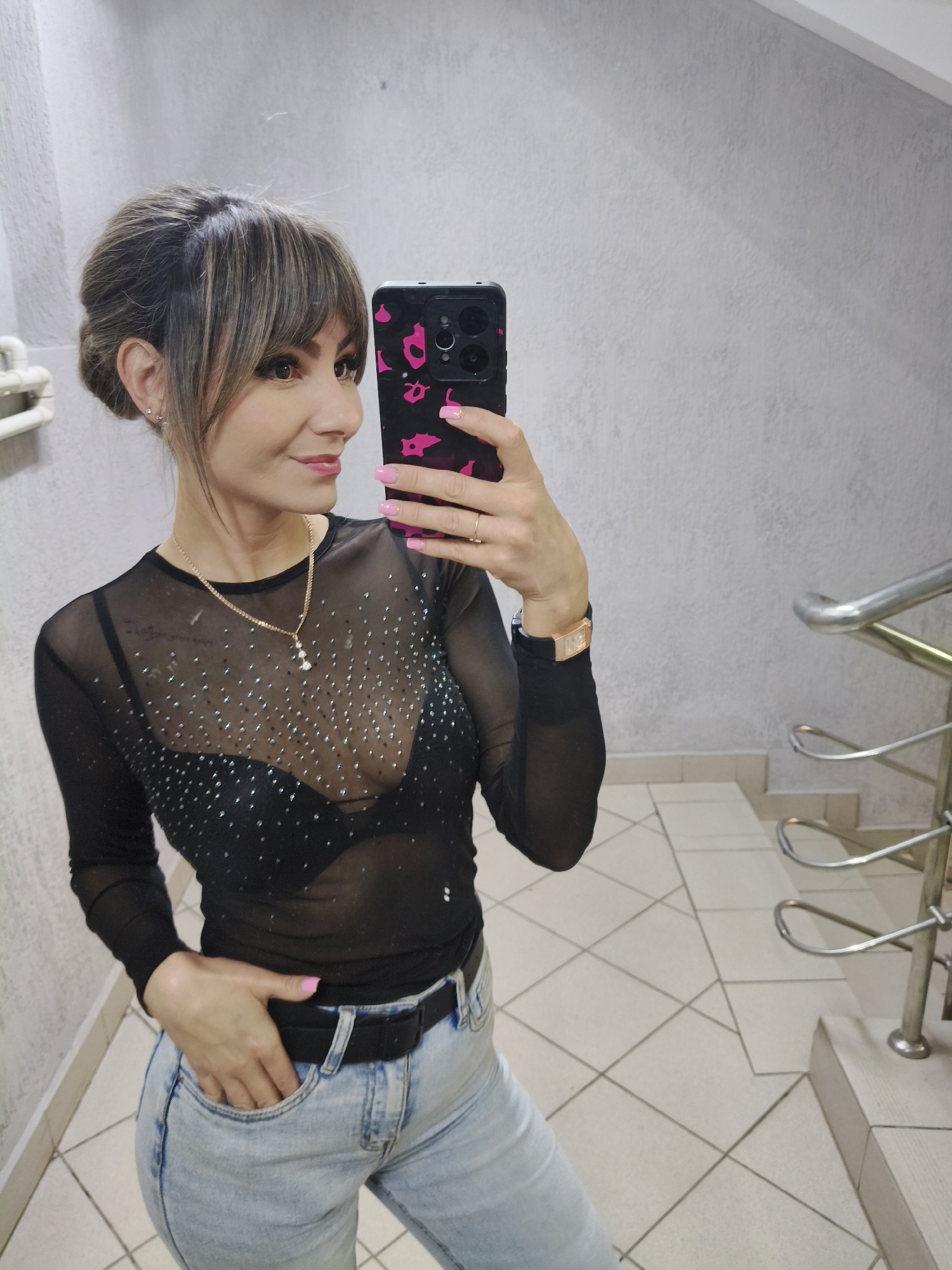 A woman is captured in a mirror selfie, posing indoors in what appears to be a stairwell or hallway in Chaikovsky, Russia. She is seen from the waist up, holding a smartphone with a black and pink patterned case in her right hand to take the photograph. Her hair is styled in an elegant updo with bangs, framing her face, which features a soft, pleasant smile and light makeup.
She is wearing a sheer black long-sleeved top, adorned with numerous scattered rhinestones, revealing a black bra underneath. Her outfit is completed with light-wash blue jeans and a black belt, with her left hand casually tucked into a front pocket. Notable accessories include a gold-colored necklace with two small pendants, small stud earrings, and a watch with a rectangular gold-colored face and a black strap on her left wrist. Her fingernails are painted a bright pink.
The background consists of textured light grey walls, a tiled floor with light-colored square tiles, and a metal handrail with looped supports on the right side, indicating a staircase. In the upper left, a white pipe or conduit is visible on the wall. The lighting suggests an indoor setting, and the woman projects an air of confidence and relaxed elegance.