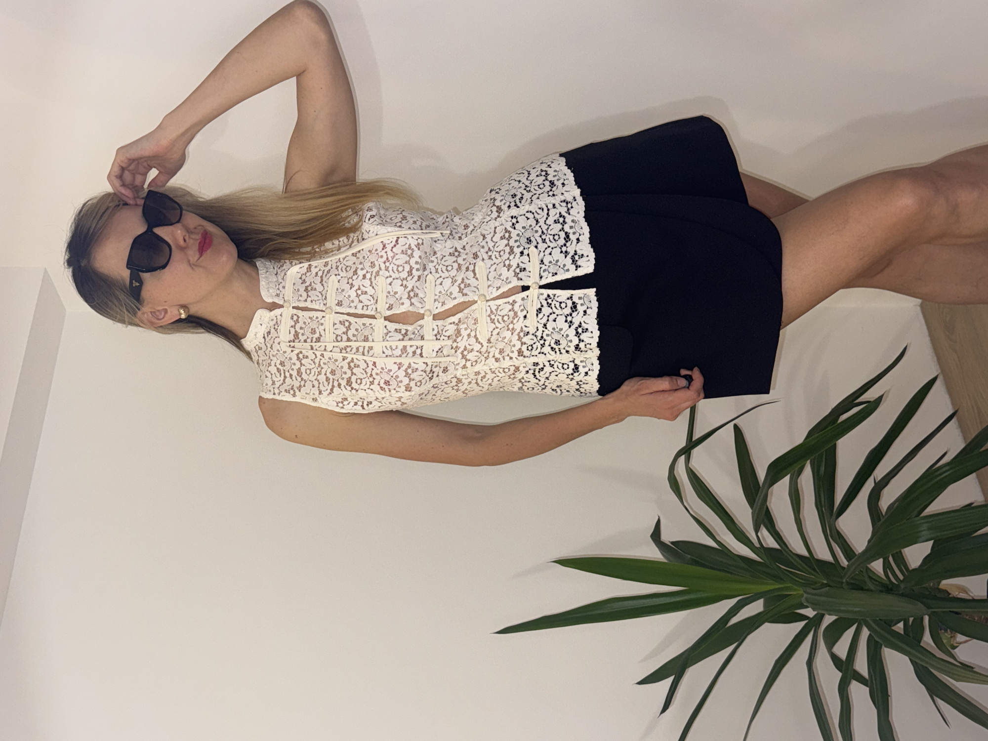 A woman is reclining, possibly posing, against a white wall. She wears dark sunglasses, a sleeveless, lacy, white top with a Mandarin collar, and a short, dark skirt. Her left arm is raised, holding the sunglasses. A potted plant with long, green leaves is in the foreground. The location is not specified in the image. Thus, I am unable to provide the location in the city of Carnikava, Latvia.