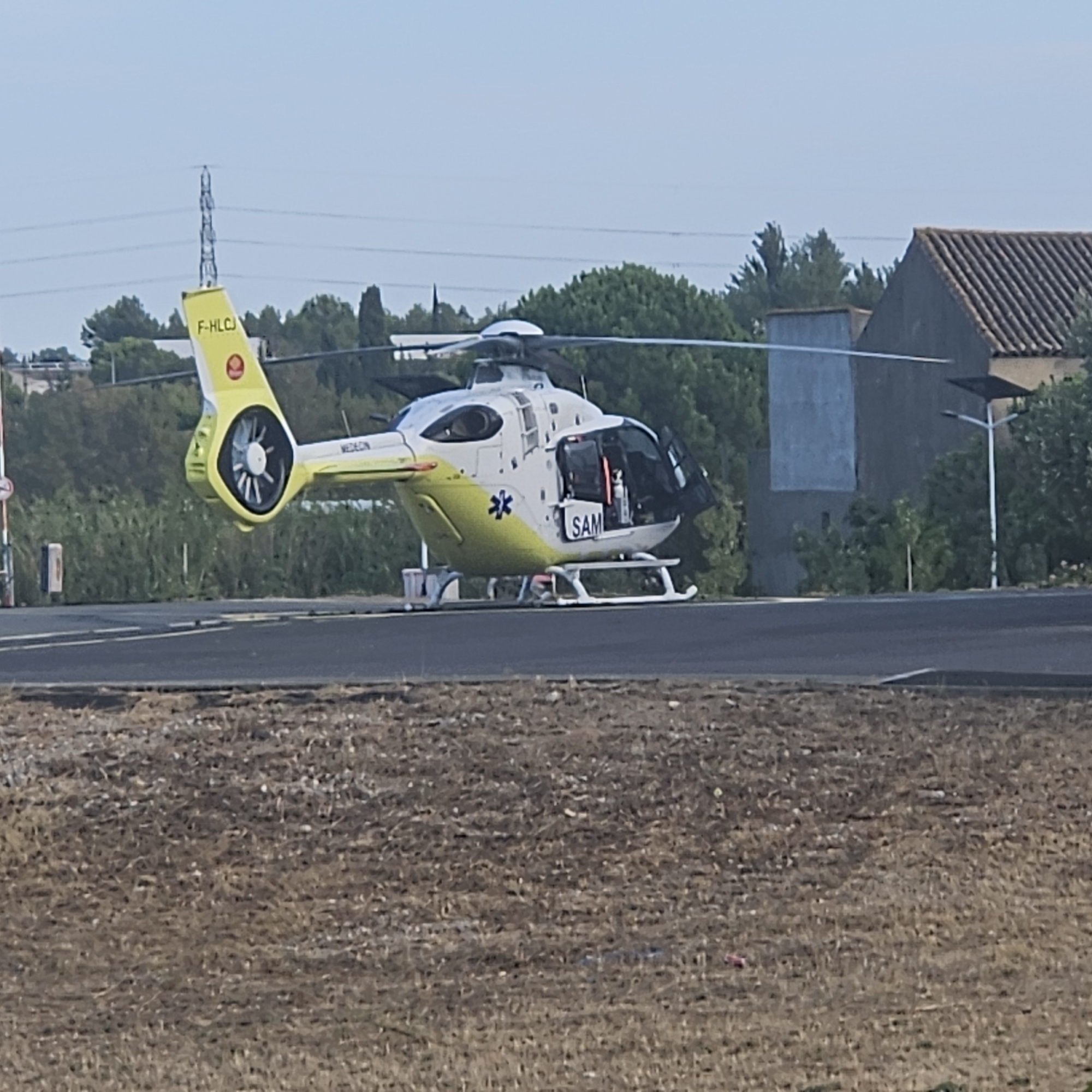 An aerial view of a white and yellow helicopter with the registration F-HLJC is parked on an asphalt pad. The helicopter has its doors open, revealing medical equipment inside. A blue star symbol and the word "SAM" are visible on the side of the helicopter. The helicopter is in a field with trees and buildings in the background. The location is the Narbonne hospital in Narbonne, France. The helicopter appears to be a medical evacuation helicopter.