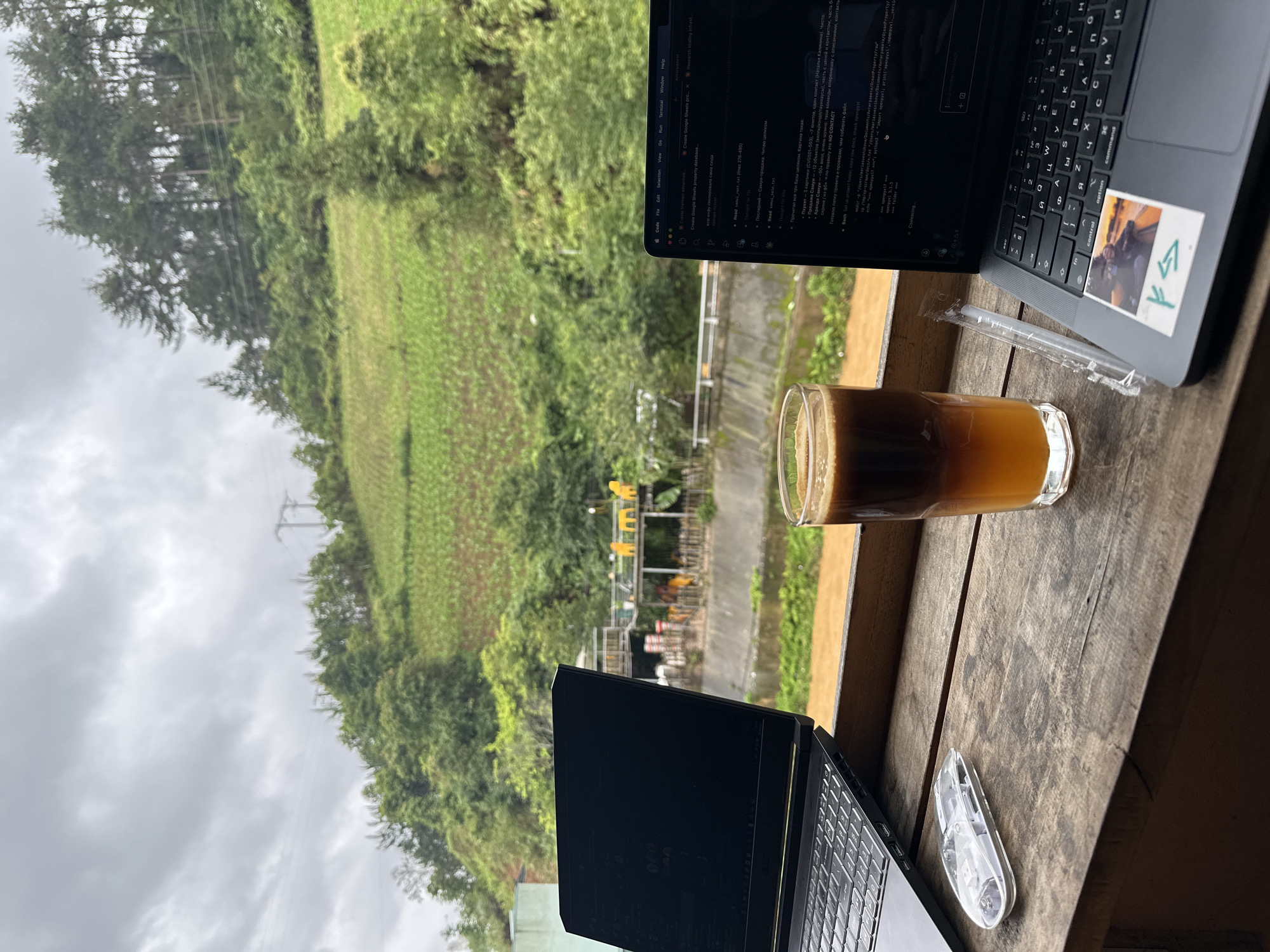 The image provides an overhead perspective from a wooden surface, overlooking a lush, mountainous landscape typical of Mù Cang Chải, Vietnam, under an overcast sky.
In the foreground, a rustic wooden table holds two open laptops and a clear glass of iced coffee, layered with dark coffee on the bottom and a lighter, frothy top. A plastic-wrapped straw lies beside the glass. The laptop on the lower left is dark-colored, while the laptop on the upper right is a MacBook, featuring a sticker on its palm rest with a small image of a person and a "K4" symbol. A small, clear, possibly a USB drive, is placed between the bottom laptop and the coffee.
Both laptop screens display text editors or code environments. The MacBook screen shows menu options ("Code", "File", "Edit", "Selection", "View", "Go", "Run", "Terminal", "Window", "Help") and several open tabs or files. Visible text includes "Create Vietnam adventure," "Create Google Sheets propert," "Research scalling infrast...", "sanul_rent.txt," "samui_sale.txt," and "openpyxl." A significant amount of Russian or Cyrillic text is present on the MacBook screen and keyboard keys, referencing "Пхукет" (Phuket), "Продажа Самуи" (Samui sales), "Аренда Самуи" (Samui rent), and the phrase "NO CONTACT."
Beyond the table, the view extends over a valley covered in vibrant green terraced fields, likely rice paddies, interspersed with trees. Power lines are visible stretching across the landscape. In the distant mid-ground, faint yellow structures can be seen. The scene depicts a tranquil, outdoor workspace, combining remote work with natural scenery.