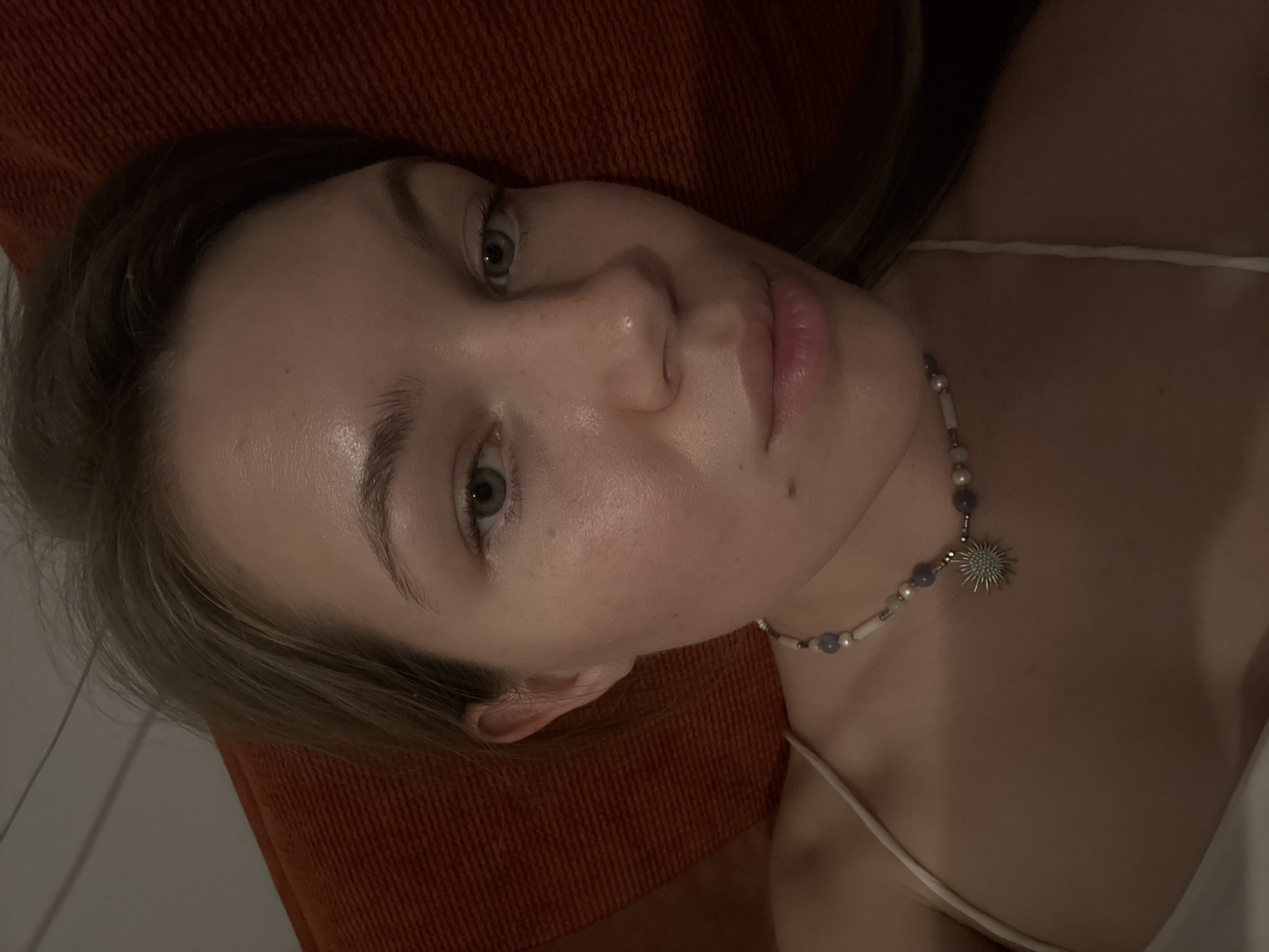 A close-up, slightly rotated, indoor photograph captures a fair-skinned woman with light-colored hair, a necklace with a sun-like pendant, and a spaghetti-strap top. She is lying against an orange, textured surface. The location is unknown and there is no indication it is in the city of Vozdovac Urban Municipality, Serbia.