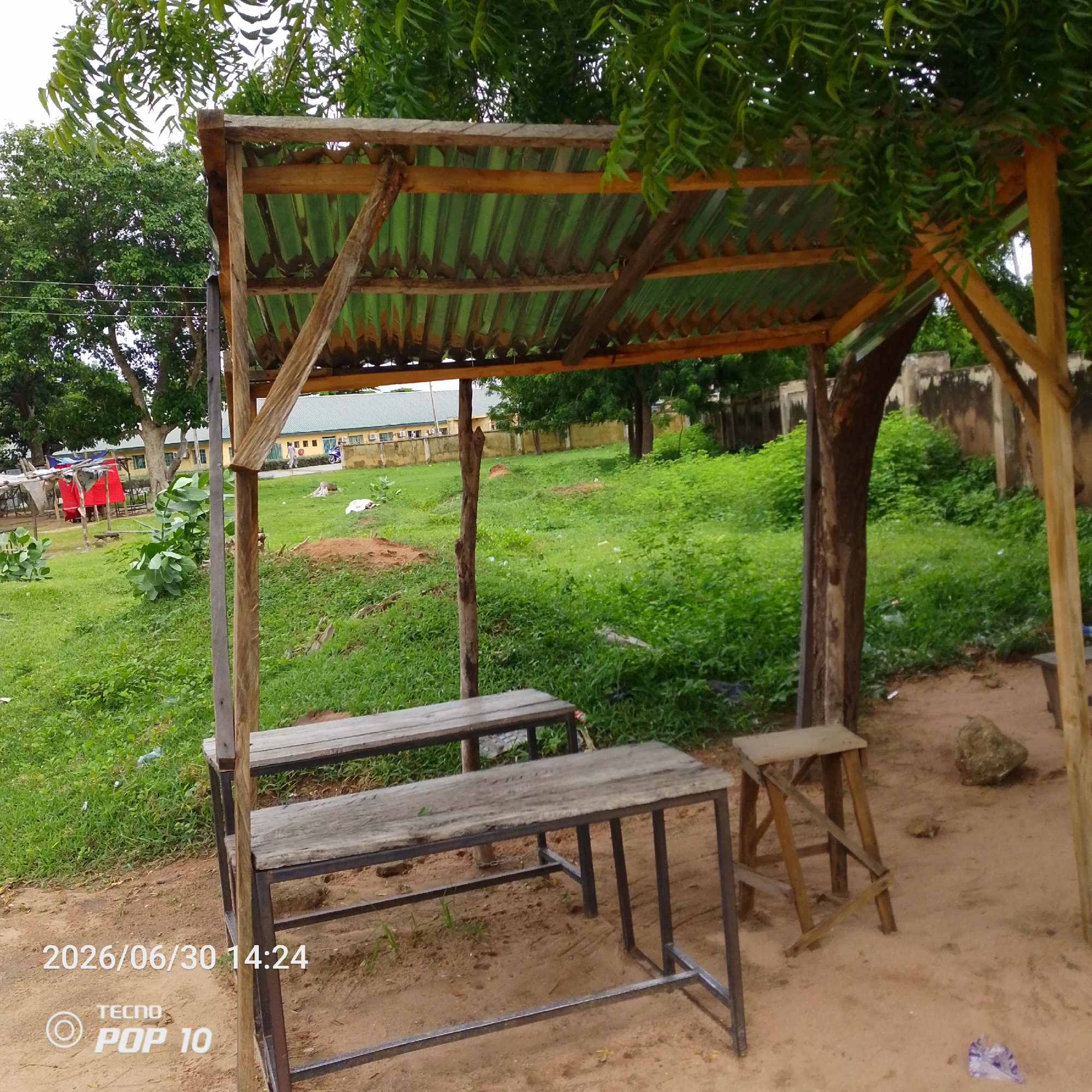 This image captures a simple outdoor shelter in Jalingo, Nigeria. The shelter, constructed with weathered wood and corrugated metal roofing, stands amidst lush green vegetation and sandy ground. Two wooden benches with metal frames are positioned beneath the shelter, suggesting a place for rest or waiting. A small wooden stool is also visible to the right of the benches. In the background, a building with a yellow facade and a beige wall can be seen, indicating a school or similar institution. Several trees, including a large one with a textured trunk, are part of the scene, providing shade. The overall atmosphere is tranquil and appears to be during the daytime, with overcast skies suggesting cloudy weather. The ground is a mix of sand and grass, with some scattered debris, including a plastic bag. The timestamp "2026/06/30 14:24" and the text "TECNO POP 10" are overlaid on the bottom left corner of the image, likely indicating the date and time the photo was taken and the device used. There are no people actively engaged in any specific activity, giving the impression of a quiet, everyday scene.
