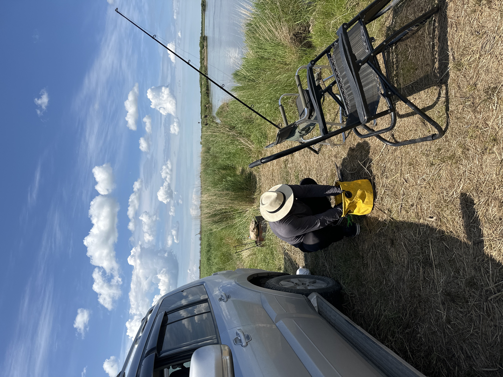 A person wearing a hat and dark clothing squats next to a silver SUV, reaching into a yellow bucket. The scene is outdoors with a body of water and tall grass in the background, under a cloudy blue sky. Two folding chairs are set up near the water. The location is identified as Im. Tel'mana, Russia.