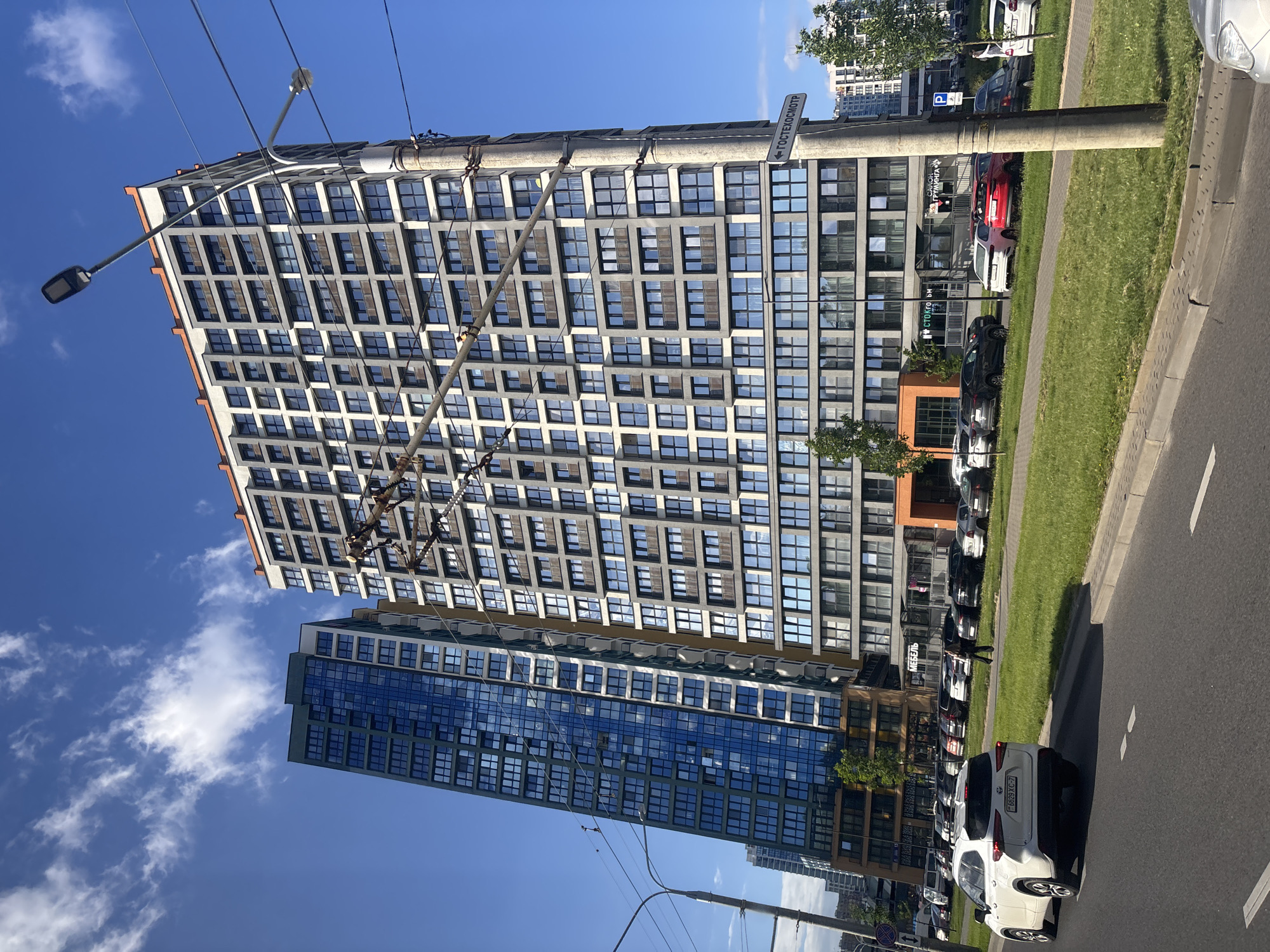 In the city of Senitsa, Belarus, a modern residential complex is captured under a bright blue sky with scattered white clouds. The complex features two prominent high-rise buildings. The building on the right is a multi-story structure with a gray facade, numerous windows, and an orange-trimmed roofline. To its left, a taller, slender building displays a striking blue glass facade. In front of the buildings, a multi-lane road is visible, with a white SUV in the foreground and several other parked cars lining the roadside. A pedestrian walks on a grassy median between the road and the buildings. Overhead, power lines and a street lamp extend across the frame, partially obscuring the buildings. A street sign with Cyrillic text, "ГОСТЕХОСМОТР," is mounted on a concrete pole. The environment appears to be an urban setting with well-maintained greenery.