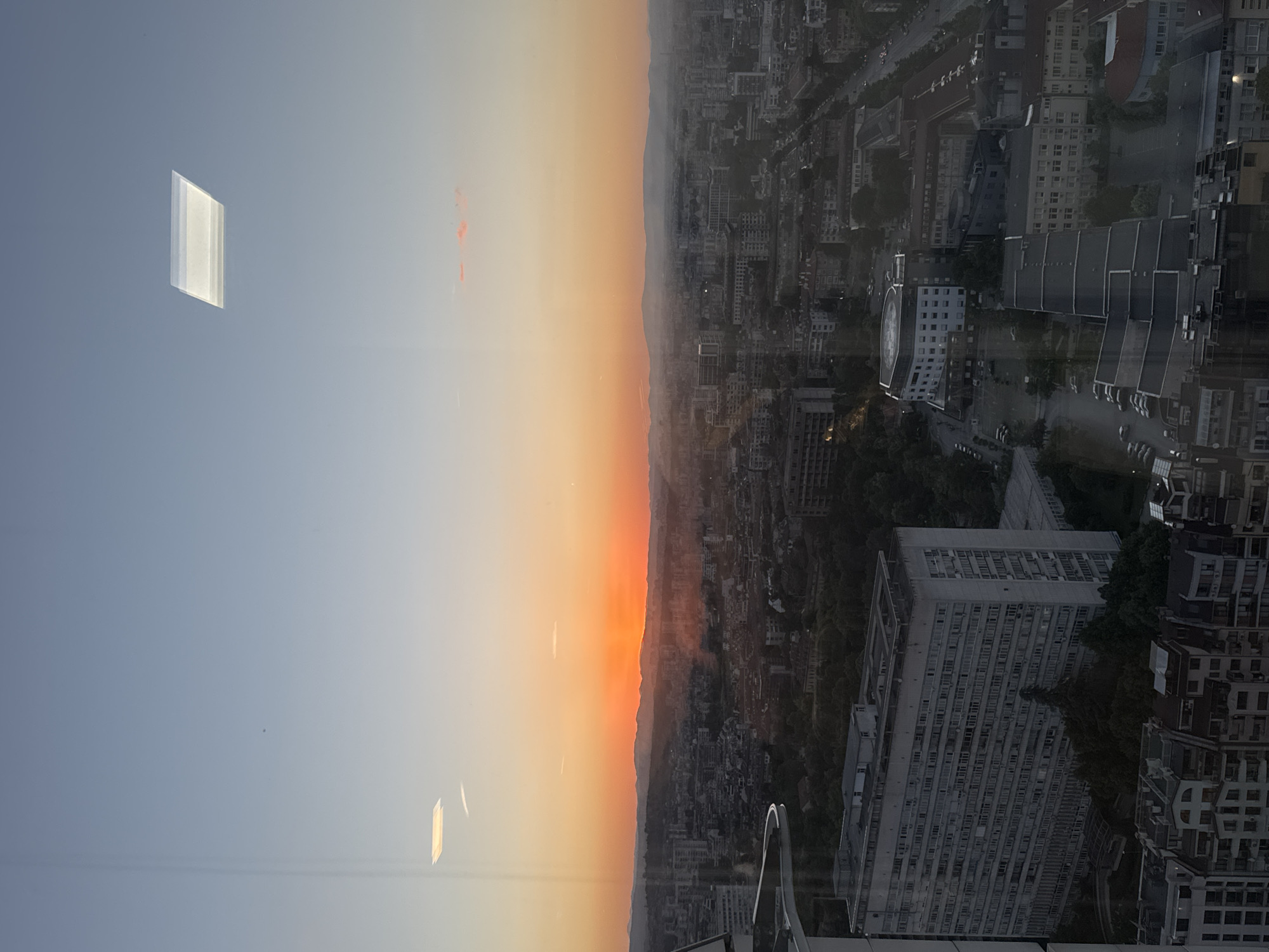 The image presents a cityscape, most likely taken from a high vantage point within a building, possibly a skyscraper. The left side of the image depicts a twilight or sunset sky gradient, transitioning from pale blue to orange near the horizon. The right side reveals a dense urban environment. A mix of building types, including high-rises and smaller structures, is visible. This is most likely Sofia, Bulgaria, based on the style of architecture. The sky is partly visible due to the building windows. An architectural detail, likely a balcony railing, appears at the bottom edge of the frame, suggesting the perspective is from inside a structure. The scene evokes a sense of tranquility juxtaposed with urban density.