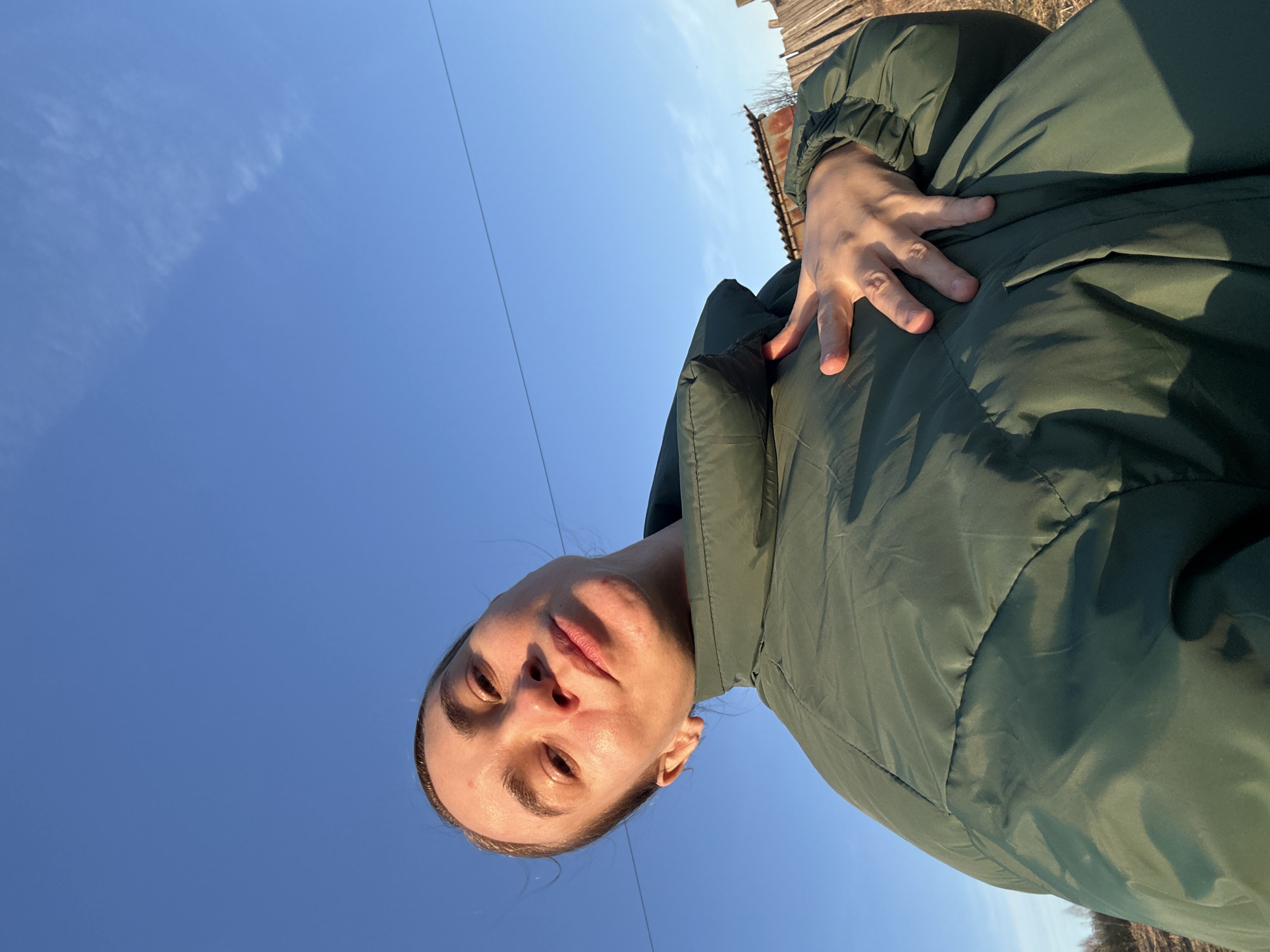 A woman is posing for a selfie outdoors. She is wearing a green padded jacket and is looking down at the camera. The background is a clear blue sky and a wooden fence or structure. The location is not evident in the image and there is no way to determine if it is Kharnavy, Russia.