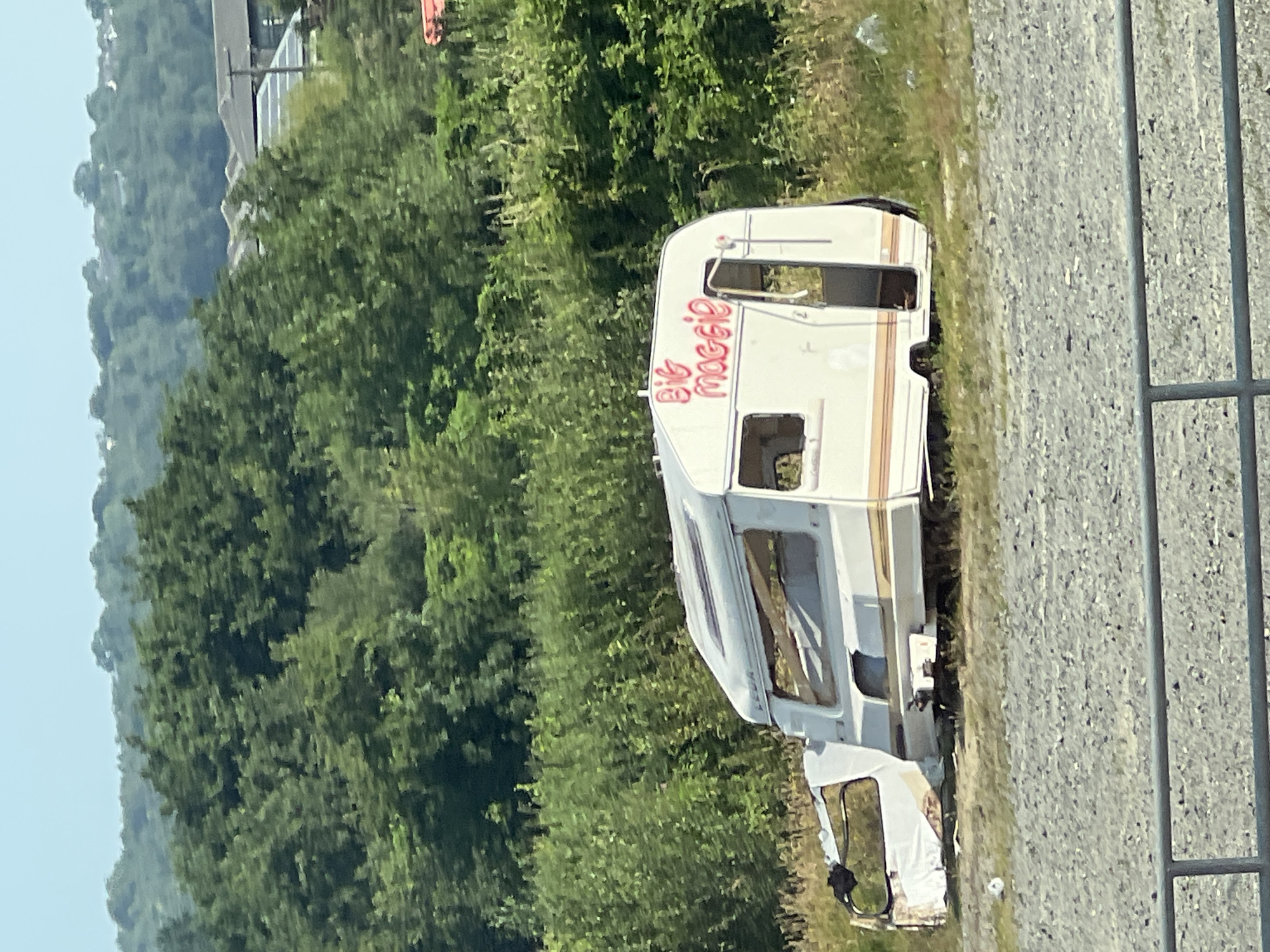 An abandoned caravan with the words "Big Maggie" spray-painted in pink on its side sits in a gravel lot. The caravan is white with orange and brown stripes. The front of the caravan appears to be damaged and is covered with white sheeting. In the background, a line of lush green trees forms a backdrop against a hazy sky. The overall scene suggests neglect and disuse. The location is not specified to be in Letterkenny, Ireland.