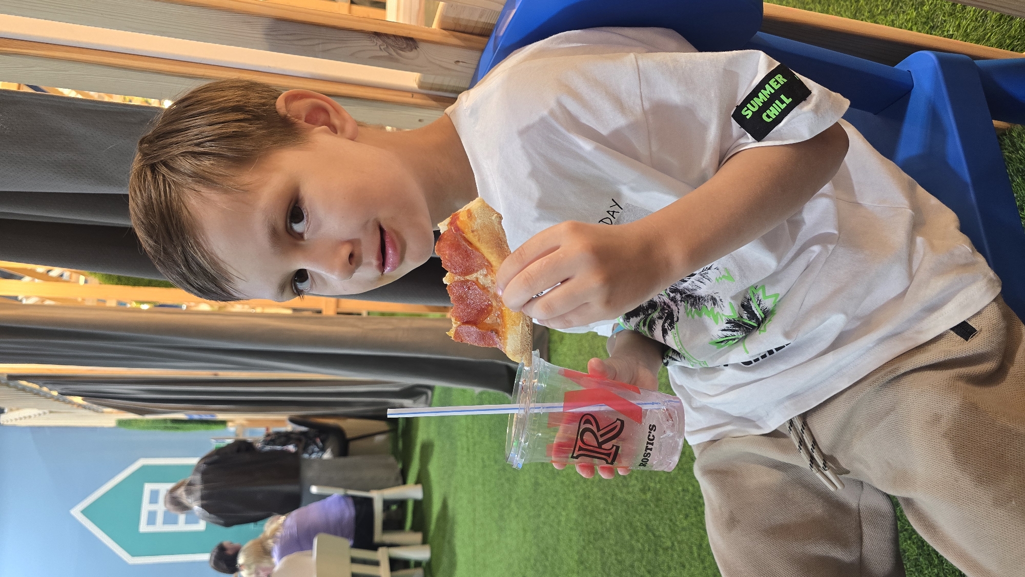 A young boy, approximately 5-8 years old, with light brown hair, is seated in a blue plastic chair. He wears a white t-shirt with a green and black graphic of palm trees on the front and a black patch on the left sleeve that reads "SUMMER CHILL" in green text. His lower attire consists of light brown sweatpants with a black and white patterned drawstring. In his right hand, he holds a slice of pepperoni pizza, distinct for its heart-shaped pepperoni toppings. His left hand grasps a clear plastic cup containing a beverage with ice and a blue and white straw. The cup displays a red stylized "R" logo above the word "ROSTIC'S". The boy is looking directly forward, towards the viewer, with his mouth slightly open.
The immediate environment features a floor covered in green artificial turf. In the background, grey curtains hang alongside wooden vertical slats or beams. Further to the left, a light blue wall is visible, adorned with a white outline of a house including windows. Several other individuals are present in the blurred background; some are seated at white tables and chairs, while one person in a dark jacket stands near the blue wall with the house outline. The setting is an indoor establishment, likely a fast-food restaurant or family-friendly dining area, identifiable as a Rostik's establishment in Izhevsk, Russia, based on the branding on the cup.