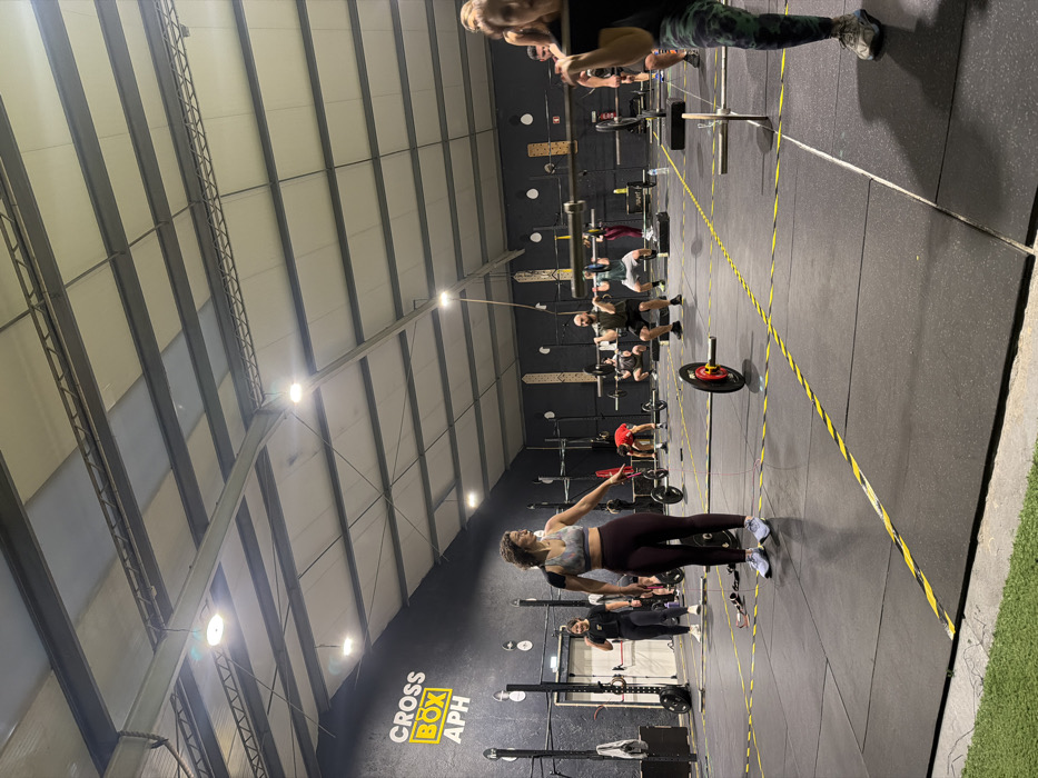 Inside a gym, individuals exercise in a structured setting. Some participants are engaged in weightlifting near squat racks marked by yellow lines. A woman in sportswear appears to be demonstrating or warming up, with her arm extended. The gym has a high ceiling with visible metal beams and bright lighting, contrasting against the dark wall with "CROSS BOX APH" printed on it. The floor is covered in dark rubber mats.
