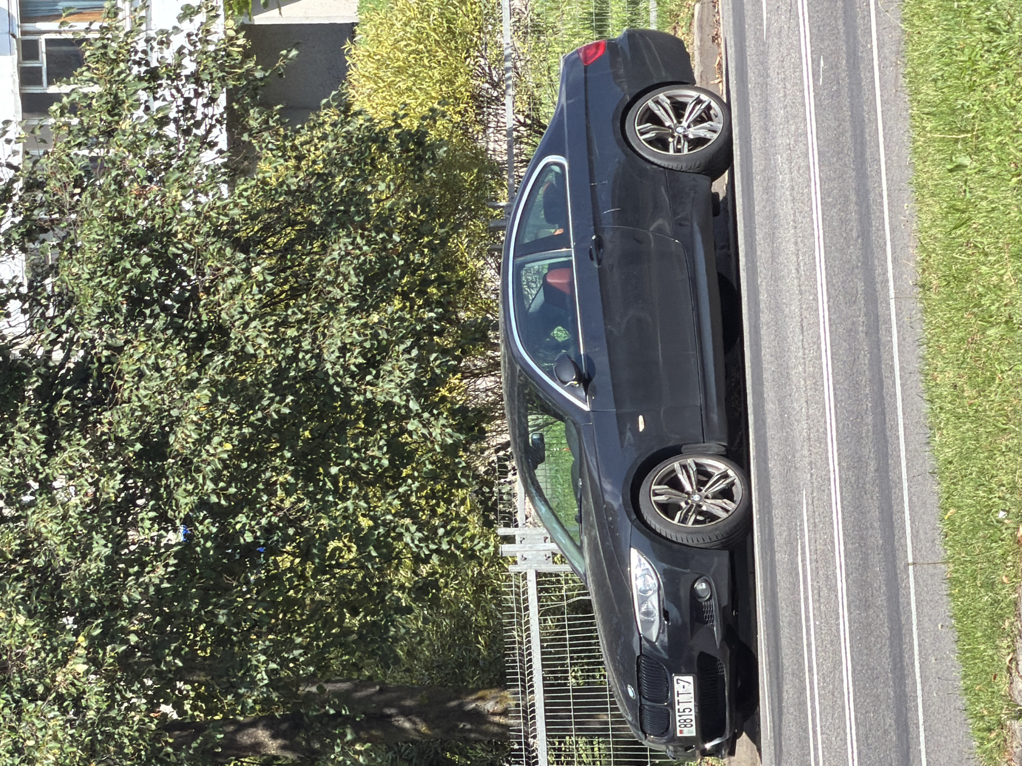 A dark-colored two-door BMW coupe with a red interior is parked on a city street in Minsk, Belarus, on a sunny day. The car is positioned on the right side of the frame, facing left. It has a Belarus license plate displaying "8815 TT-7". A metal fence runs horizontally behind the car, separating it from a sidewalk and a line of trees and bushes. In the background, a portion of a multi-story building is visible through the foliage. The street in front of the car has white lane markings.