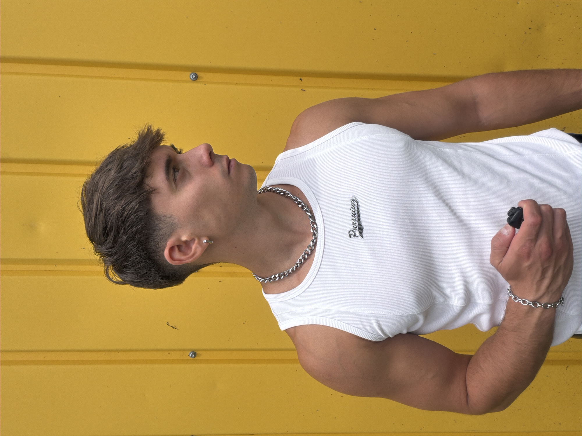 A young man with brown hair and fair skin is captured in a portrait against a bright yellow background. He is wearing a white ribbed tank top, a silver chain necklace, and a silver bracelet. His left arm is bent, and he is holding a black object in his hand. The man has a silver hoop earring in his left ear. The yellow background appears to be a corrugated metal surface, with visible screws or rivets. The lighting suggests it is daytime, likely outdoors, with clear weather. The man's gaze is directed upwards and to his left, giving him a thoughtful or contemplative expression. The word "Pursuit" is embroidered in black text on his tank top. The image is oriented vertically. The location is Sofia, Bulgaria.