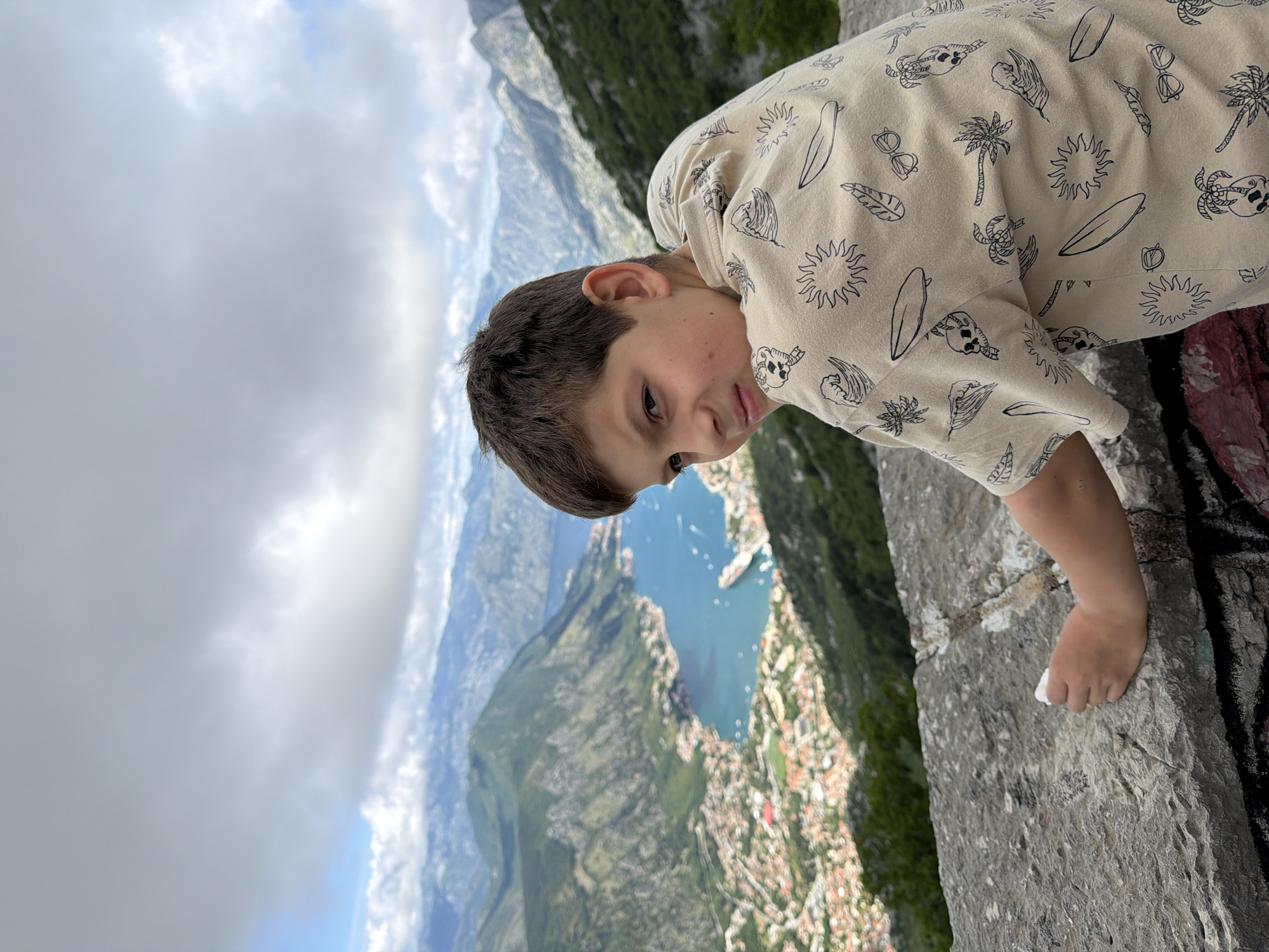 A young boy with dark brown hair and dark eyes is captured leaning over a weathered stone wall, looking downwards and sideways into a vast landscape. The image is rotated, showing the boy's head tilted towards the camera with a playful expression, including a slight smirk and his tongue peeking out. He wears a light-colored, long-sleeved hooded shirt adorned with black outline patterns of suns, palm trees, surfboards, sunglasses, and skulls. His right hand rests casually on the stone wall.
Beyond the boy and the foreground wall, a stunning panoramic view unfolds. A large, deep blue body of water, consistent with a bay or fjord, is visible, reflecting the partially cloudy sky. Along the shores of the water, a town with numerous buildings, some featuring prominent red roofs, is nestled, extending up the surrounding green, steep mountainsides. Several small boats dot the water. The sky shows a mix of white clouds and brighter areas where sunlight is breaking through, suggesting it is daytime. The scene strongly depicts the natural beauty of a coastal mountainous region, specifically aligning with the panoramic views found in Špiljari, Montenegro, overlooking the Bay of Kotor. The boy's pose and expression convey a sense of curiosity and engagement with the impressive scenery below.