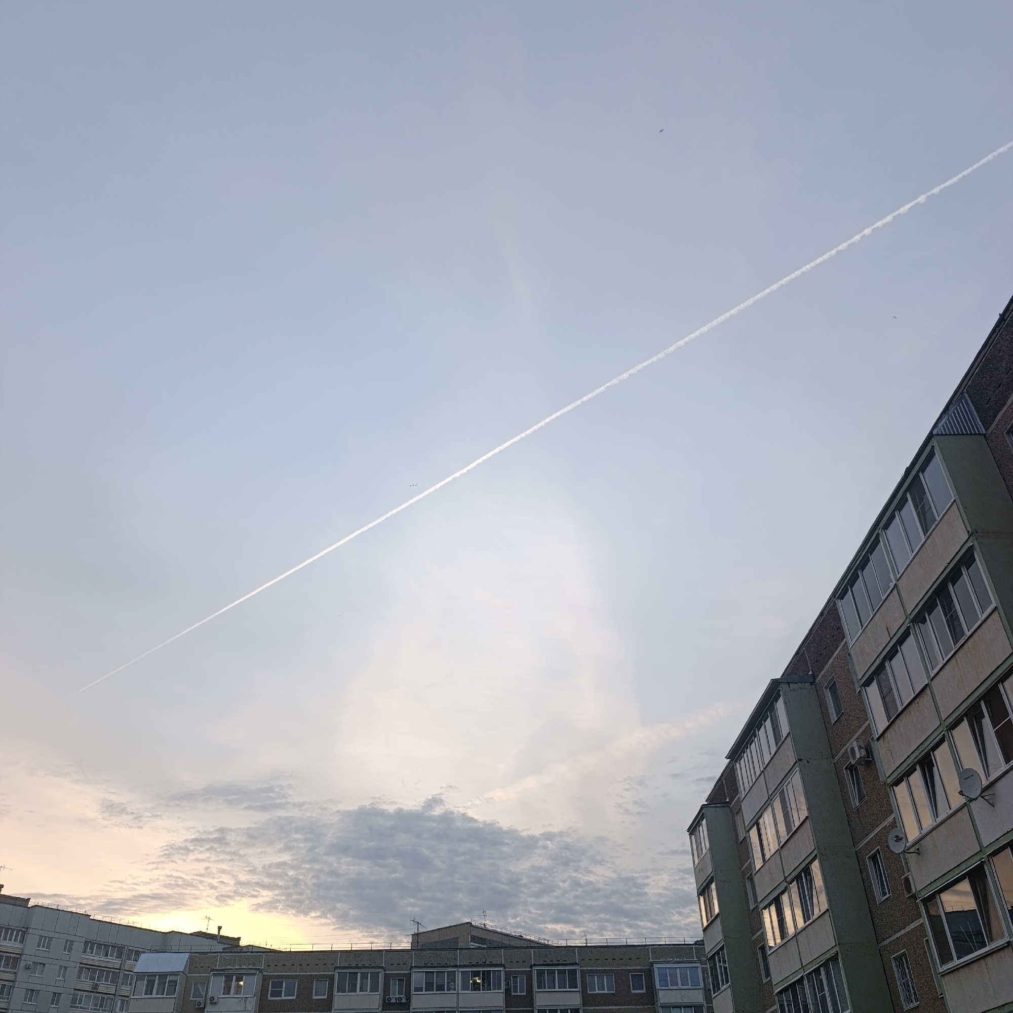 The image depicts a view of apartment buildings in the city of Chaikovsky, Russia, under a cloudy sky with a visible contrail. The buildings are multi-story and appear to be constructed of concrete with numerous windows and balconies. The sky is overcast with some lighter patches near the horizon. A distinct white contrail stretches diagonally across the upper portion of the frame. A small bird is also visible in the sky.