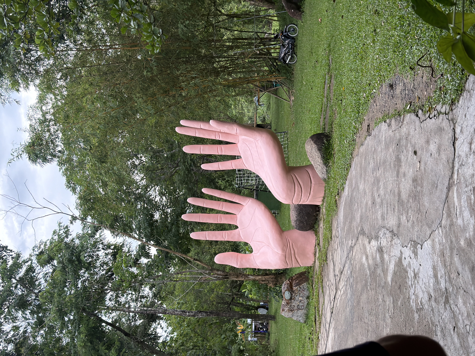 In Khanh Vinh, Vietnam, a park features a concrete path winding through lush green grass and trees. Two enormous, light pink sculpted hands with fingers extended upwards are prominently displayed on the grass, positioned to the right of the path. Large, rounded boulders are placed around the base of each hand. In the background, a dark-colored motorcycle is parked on the grass. The environment is a natural landscape with bamboo trees and other foliage.