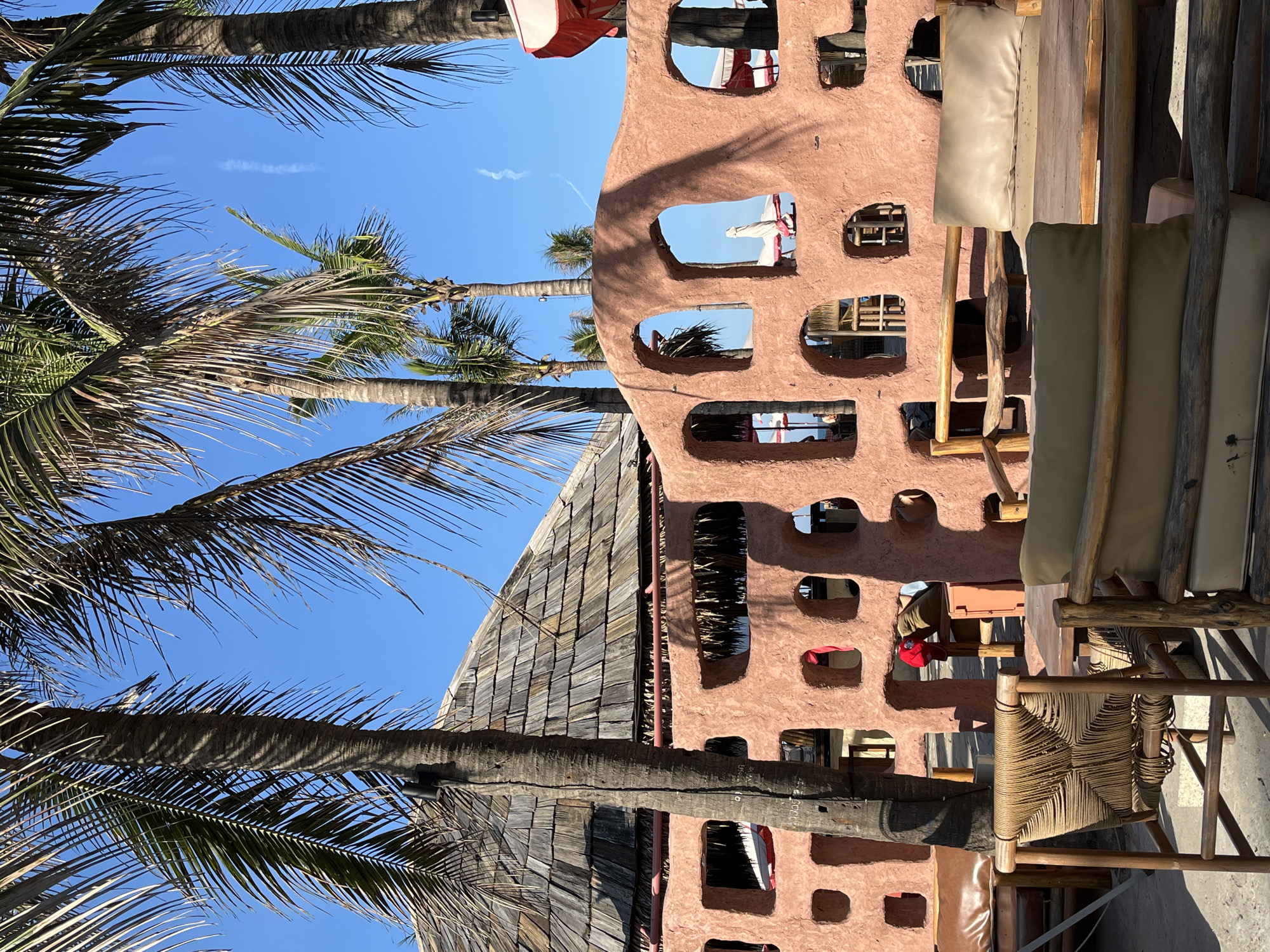 Captured from a low angle, the image presents a sun-drenched scene in Đà Nẵng, Vietnam. A vibrant pink-red building with irregularly spaced, round windows is prominently featured, offering glimpses into the interior and the outdoor scene. Palm trees, silhouetted against a clear blue sky, add a tropical ambiance. In the foreground, rustic wooden furniture, including chairs with woven backs and neutral-colored cushions, suggests a relaxed, outdoor dining setting. The image's composition and elements evoke a sense of exotic travel.