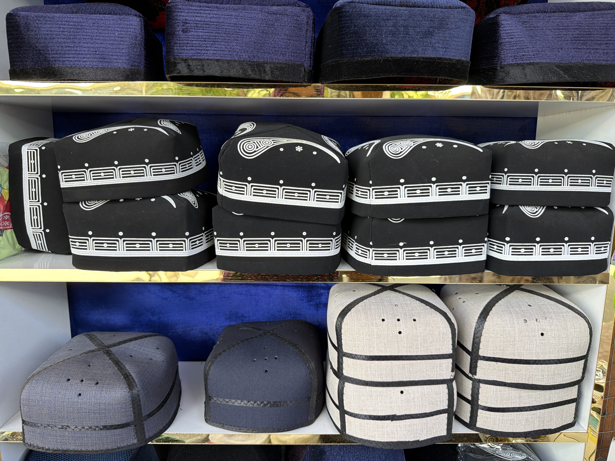 The image shows a shelf display of kufis, which are traditional skullcaps. The kufis are arranged on multiple shelves, with rows of blue kufis at the top, followed by black kufis with white embroidered patterns, and then kufis in grey, dark blue, and beige with black stitching. The display appears to be in a market or shop setting, likely in Zangiata, Uzbekistan, given the types of kufis shown. The lighting suggests an indoor environment. There are no people visible in the image.