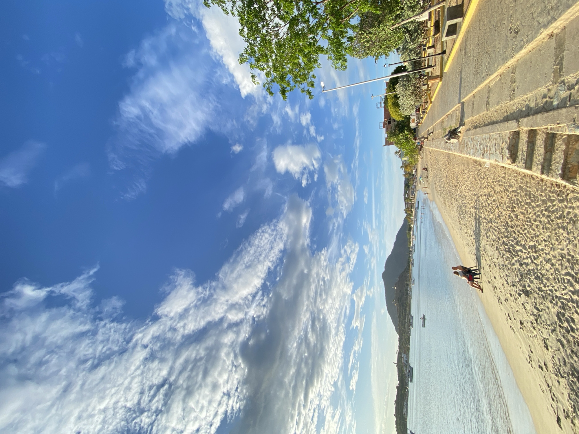 In Juan Griego, Venezuela, a beach scene unfolds with a wide expanse of sand meeting the tranquil waters of the Caribbean Sea. The sky is a vibrant blue, dotted with wispy white clouds that create a sense of depth and movement. In the distance, a prominent mountain looms, crowned with what appears to be a fortification or historical structure. The shoreline is lined with a paved promenade, featuring benches, lampposts, and lush green trees providing shade. Several boats are anchored in the bay, suggesting a harbor or a popular nautical area. On the sand, two individuals are seated in chairs, facing the sea, engrossed in each other's company. Further down the beach, other beachgoers can be seen scattered, some relaxing on the sand, while a lone figure walks along the water's edge. A dog is visible on the promenade, lying down near a stone wall. The overall atmosphere is peaceful and idyllic, capturing a typical day at this coastal Venezuelan town.