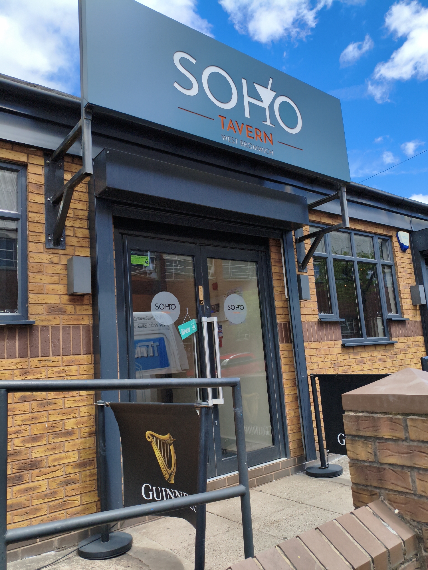 This is a medium-shot, eye-level photograph of the exterior of a pub called "Soho Tavern" located in West Bromwich, Sandwell, United Kingdom. The pub has a distinctive yellow brick facade. Above the entrance, a prominent light blue sign displays "SOHO TAVERN" in white lettering, with "WEST BROMWICH" below it.
The entrance to the pub features a dark-colored door with glass panels. A small green sign on the door reads "OPEN." Inside the glass, a white circular sign with "SOHO" is visible. To the left of the entrance, a window is partially obscured by blinds.
In front of the pub, a black barrier with chrome poles encloses a small outdoor seating area. A black banner displaying the Guinness logo and "GUINNESS" is positioned behind the barrier. The ground in front of the pub is paved with concrete.
The sky above is a vibrant blue with fluffy white clouds, suggesting it is daytime with pleasant weather. The overall impression is of a well-maintained and inviting local pub. There are no people visible in the image.