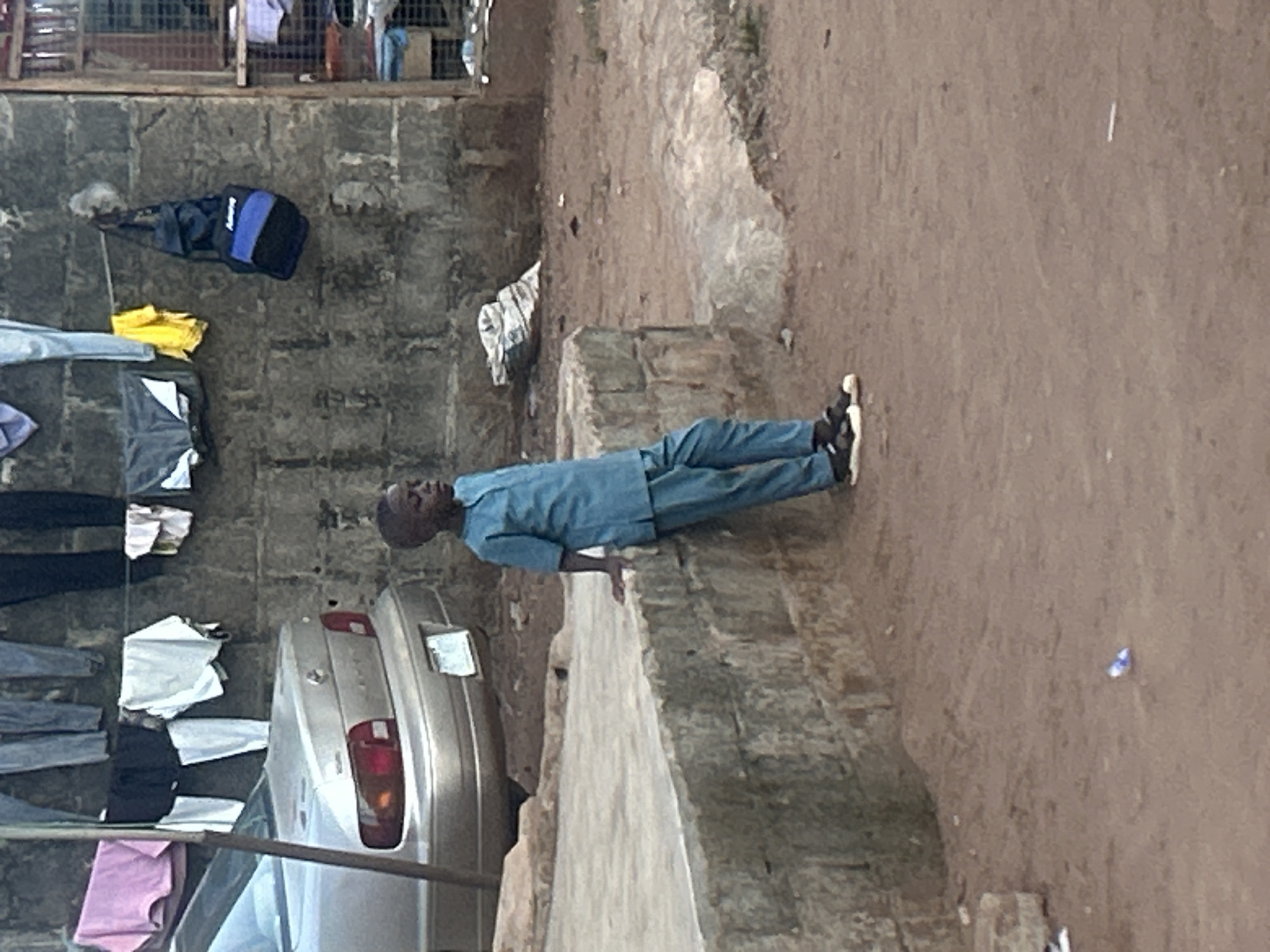 The image shows a man in a light blue outfit walking alongside a concrete wall next to a silver car. The environment appears to be an outdoor space in the city of Unknown, Unknown, with a brick wall and hanging clothes on the left side of the image. The ground is dirt.
