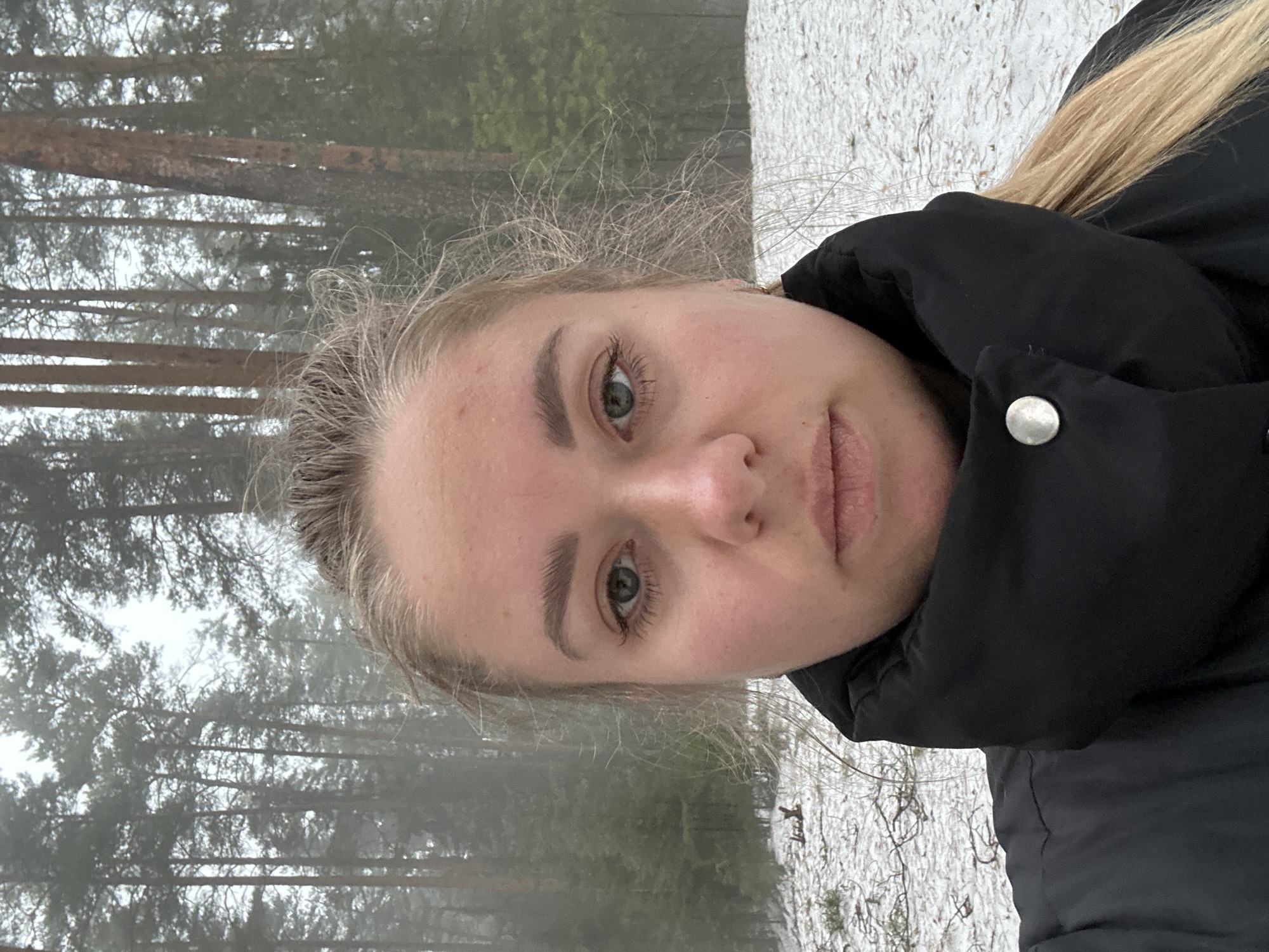 A close-up portrait of a young, light-skinned woman with blonde hair pulled back. She is wearing a black, puffy jacket with a visible silver button. The background features a forest in a somewhat foggy or overcast condition. Patches of snow are visible on the ground. Based on the location information, the scene is likely in or near the city of Chaikovsky, Russia.