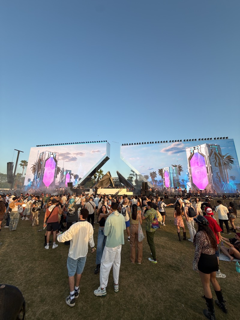 The image shows a large crowd of people gathered at an outdoor festival in Indio, United States. The crowd is standing on a grassy field with a large stage in the background. The stage is decorated with screens displaying abstract imagery and palm trees. The sky is clear and blue. The crowd appears to be a mix of ages and ethnicities, dressed in a variety of casual clothing. Some people are standing and talking, while others are moving through the crowd.