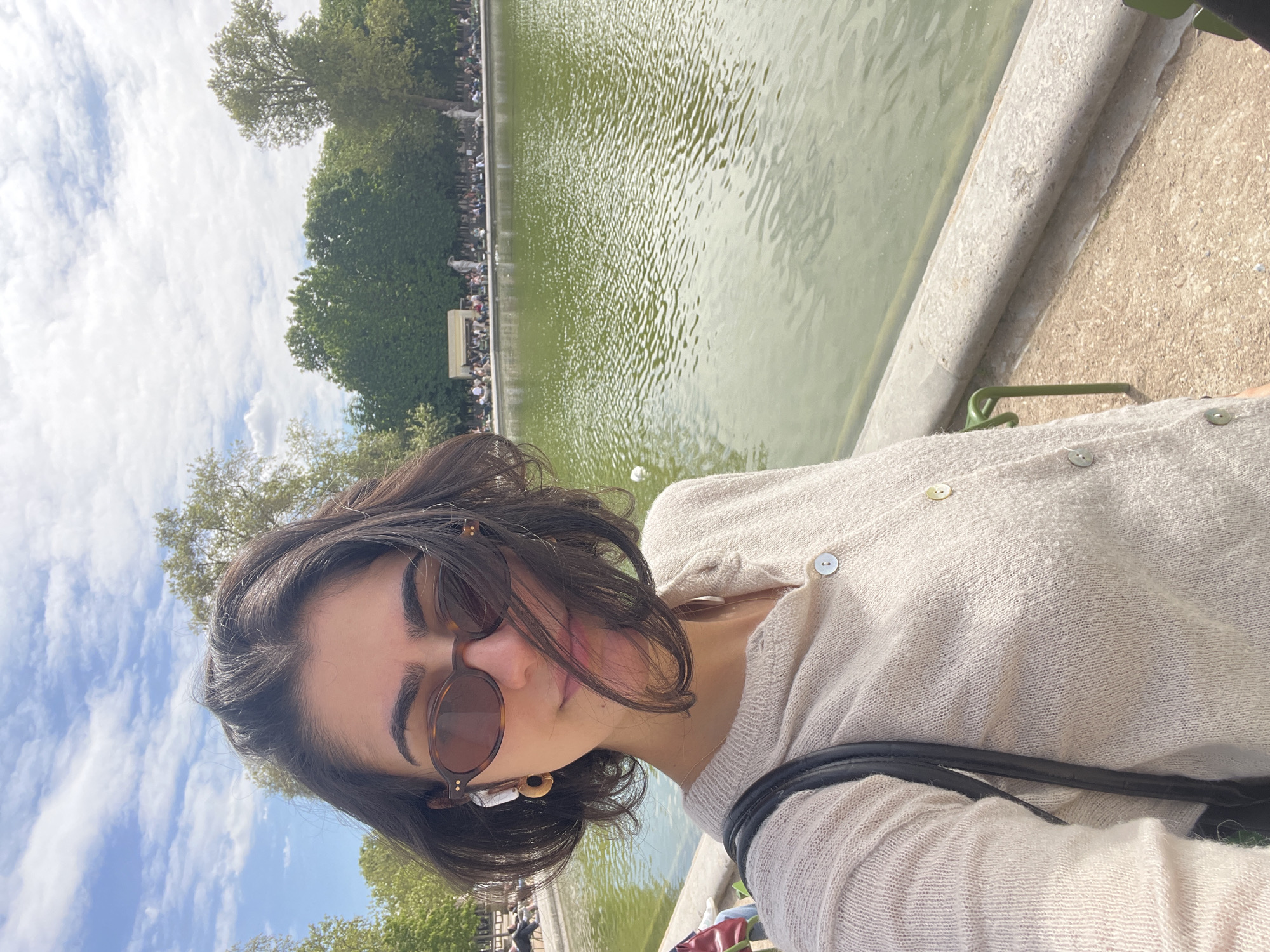 A young woman takes a selfie in a sunny outdoor setting, appearing to be in a park. She is positioned to the right, looking down slightly with a neutral expression. She has short, dark hair and wears round, tortoiseshell-patterned sunglasses. Her prominent eyebrows are visible above the frames. She is dressed in a light beige or off-white knitted cardigan with light-colored buttons and a black strap, likely from a bag, visible across her chest. A distinctive earring, with a white and brown/gold design, is visible on her right ear.
In the background, a large body of greenish water, resembling a pond or lake, stretches across the left and central parts of the frame, showing ripples and reflections. Along its far edge, numerous people are gathered, many seated, appearing to relax and enjoy the environment. Behind the crowd, a dense line of vibrant green trees frames the scene. Above the trees, a bright blue sky with scattered white clouds indicates clear, pleasant weather. The immediate foreground features a textured concrete or stone barrier bordering the water, with a small green metal fixture attached.
The image captures a leisurely daytime scene in what appears to be a public garden or park, likely in Paris, France, given the typical setup of large ponds surrounded by people in its famous parks. There is no visible text in the image.