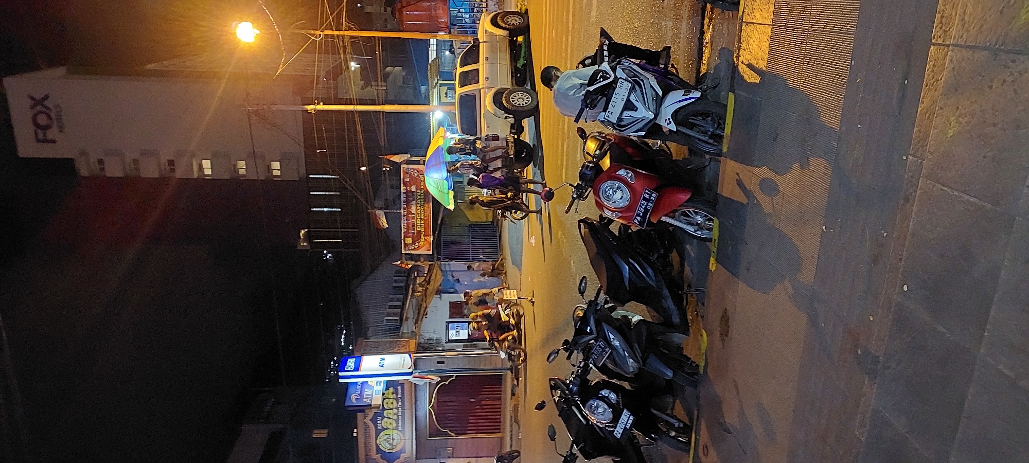 A night scene captures a street in Jayapura, Indonesia, featuring parked motorcycles and a white pickup truck. A tall, white building with "FOX HOTELS" signage stands in the background, illuminated by streetlights. In the foreground, several scooters are parked along a curb, some with yellow parking guides. A man in a light-colored shirt sits on one of the scooters. Across the street, a white pickup truck is parked with several individuals seated in its bed under a colorful umbrella. Adjacent to the truck, a sign reads "DIRGAHAYU REPUBLIK INDONESIA KE-77". A BRI ATM and a sign for "KEDAI BASK" are visible on the left side of the image. The street is paved, and shadows from the buildings and objects are cast across it.