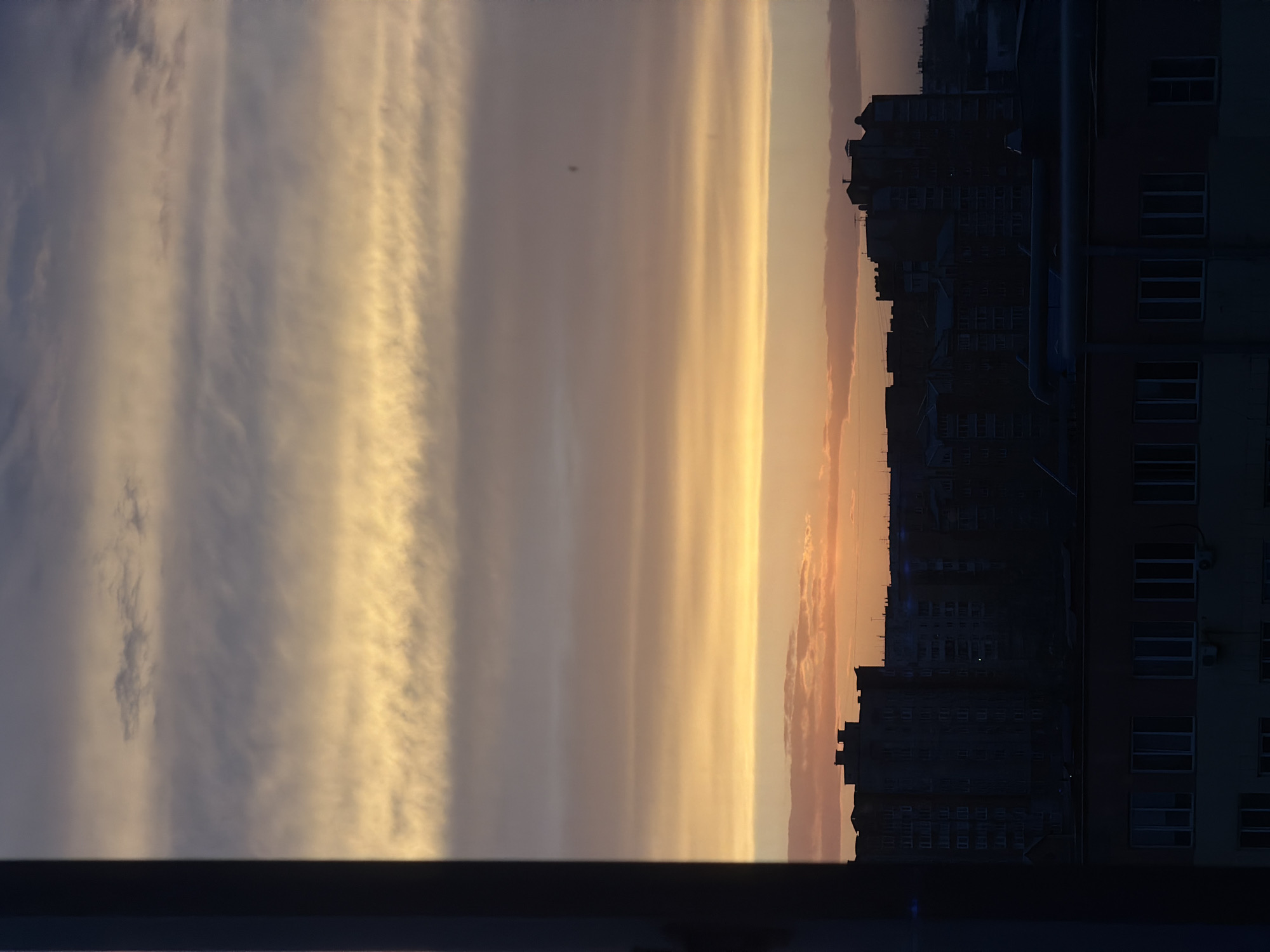 The image depicts a sunset view from a window in a city, likely Krasnoyarsk, Russia, given the urban architecture. The foreground consists of a dark windowsill. The middle ground features tall apartment buildings silhouetted against the fading sunlight. A band of orange-tinted clouds hugs the horizon. The sky above displays a striated pattern of light-colored clouds against a darker background, illuminated by the setting sun.