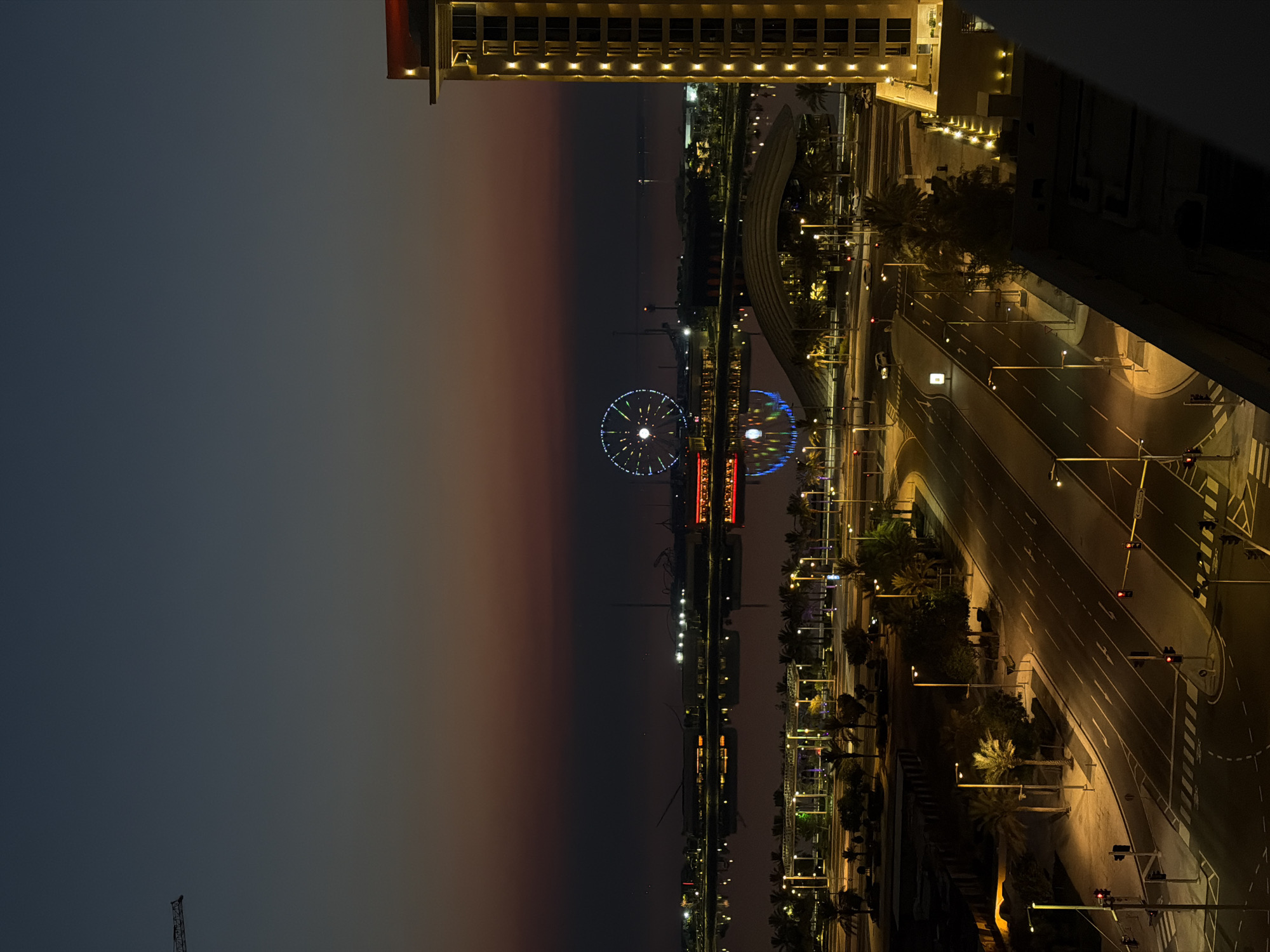 An elevated view captures a cityscape at dusk. A tall, illuminated building occupies the right side of the frame, its windows glowing with warm light. In the distance, a Ferris wheel stands out against the twilight sky, its colorful lights twinkling. The sky displays a gradient of deep blue at the top, transitioning to hues of orange and red near the horizon. A body of water reflects the city lights and the sky. A bridge arches over the water in the mid-ground, and a road with streetlights is visible in the foreground. Palm trees line the streets and walkways. The scene appears to be a developed urban area.