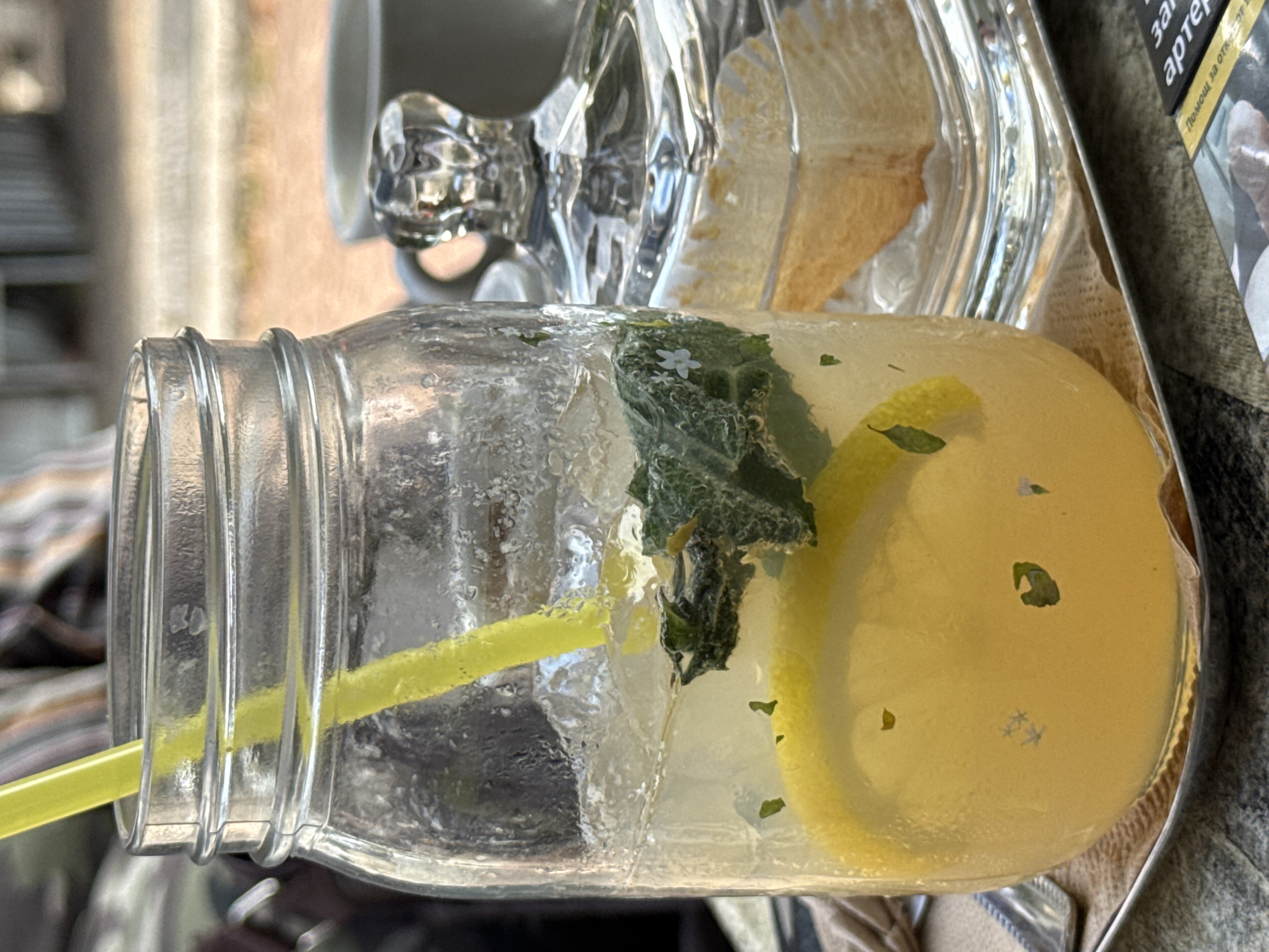 The image captures a close-up of a refreshing lemonade in a mason jar, placed on a metal tray with a napkin. The lemonade contains ice, mint leaves, and a slice of lemon. A yellow straw protrudes from the jar. In the background, a glass sugar bowl with a faceted lid sits next to a gray coffee cup. The setting appears to be an outdoor cafe, possibly in the city of Lovech, Bulgaria, given the presence of what looks like a cigarette pack with Cyrillic text in the foreground. The overall atmosphere suggests a relaxed moment, perhaps during a sunny day.