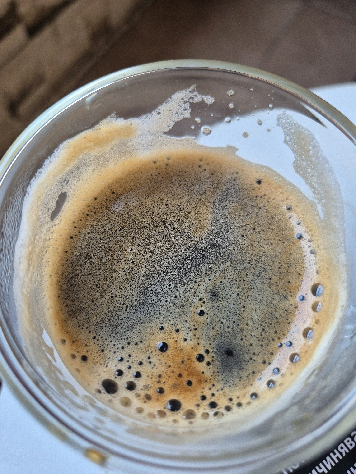 A close-up, overhead shot captures the surface of a dark, frothy espresso in a clear glass cup. The foam exhibits a range of browns, from a light tan at the edges to a deep, rich brown at the center, with numerous tiny bubbles and larger, darker ones scattered across its surface. The liquid underneath is dark and appears to have a rich crema. The glass cup is transparent, and a hint of condensation is visible on its exterior. The background is softly blurred, showing a textured, brownish surface, possibly a table or placemat, and a sliver of a white surface in the lower portion of the frame. Text is visible on the white surface, partially obscured.