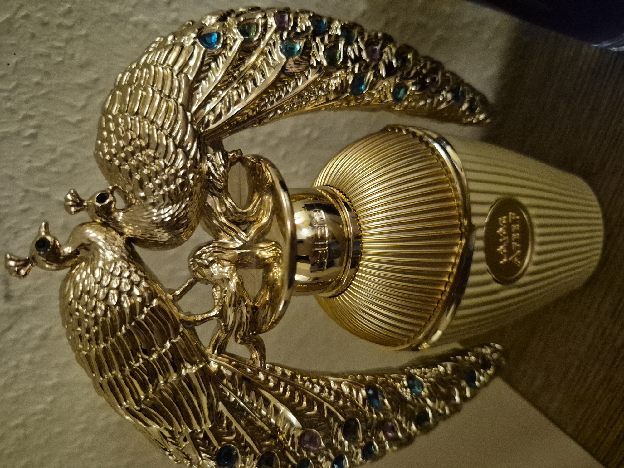 A close-up, eye-level, indoor shot shows a gold perfume bottle with two gold peacock figures adorning its top. The bottle is ribbed and stands on a flat surface, possibly a shelf. The peacocks are depicted with their tails fanned out, adorned with small, colorful gemstones in blue, green, and purple, mimicking peacock feathers. One peacock is positioned slightly in front of the other. The background is a textured, neutral-colored wall. The lighting creates a warm glow and some shadows. The scene is set in an unknown location in the city of Unknown, Unknown.