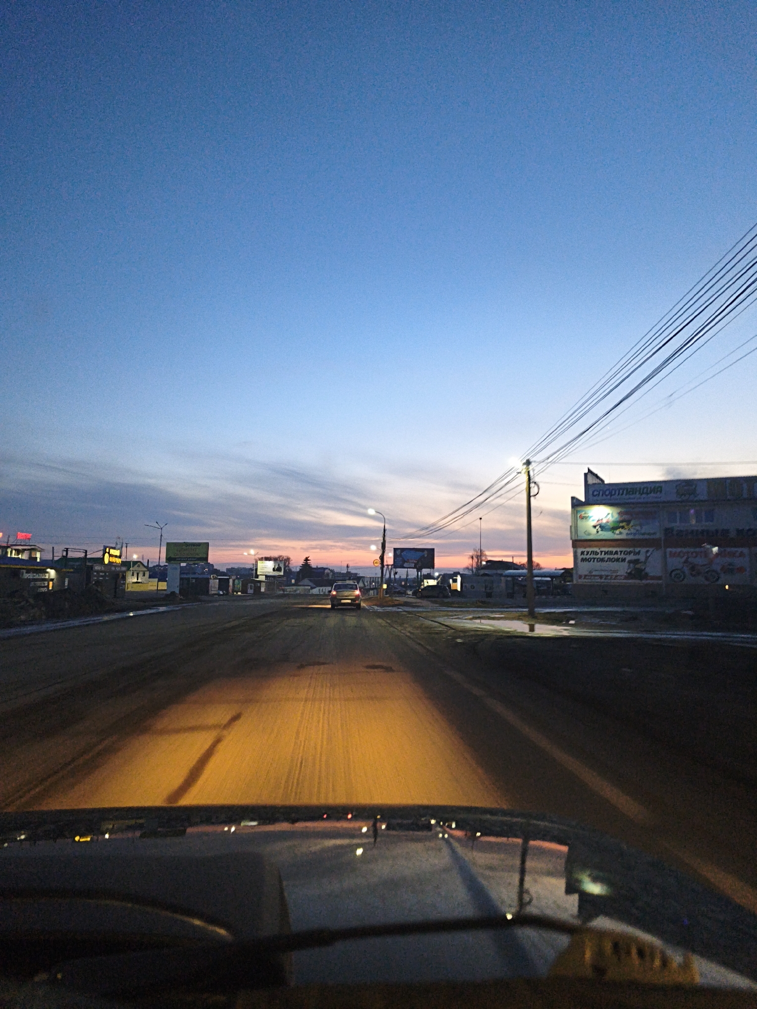 The image is a photograph taken from inside a car, looking out through the windshield at a street in Chaikovsky, Russia. The time of day appears to be early morning or late evening, judging by the state of the sky that features the hues of sunrise or sunset. The road is straight and stretches into the distance. Streetlights illuminate the road. Buildings line both sides of the street, including one prominently displaying an advertisement in Russian. Electrical lines are overhead. The car's headlights are on, illuminating the road immediately in front of it. The lower part of the windshield and the dashboard of the car are visible in the foreground.