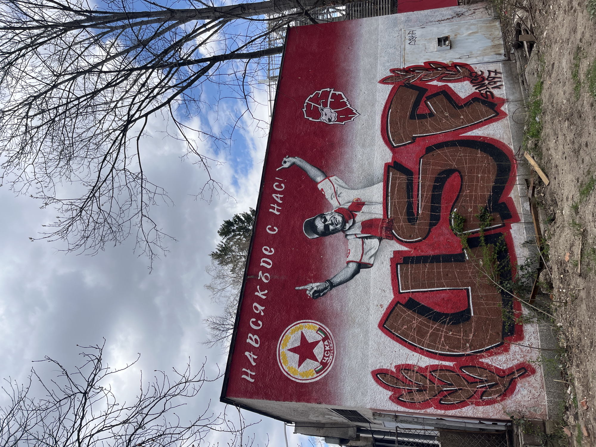 The image depicts a section of a wall mural in Sofia, Bulgaria, likely part of an outdoor art installation or public display. The mural features a color scheme of red and white, portraying a man in a sports jersey pointing, alongside the text "НАВСЯКЪДЕ С НАС!" (Everywhere with us!). There's a circular logo with a red star, yellow border, and text “ЦСКА София” (CSKA Sofia) at the base of the portrait. The artwork also includes a decorative element resembling laurel wreaths on either side of a large numeral "5," painted in brown with red outlining. The wall shows signs of wear and graffiti, with a small metallic door or vent near the upper edge. The surrounding environment includes bare tree branches against a sky with clouds and some urban elements such as parts of buildings and fencing. There is also some dirt and vegetation at the base of the wall.