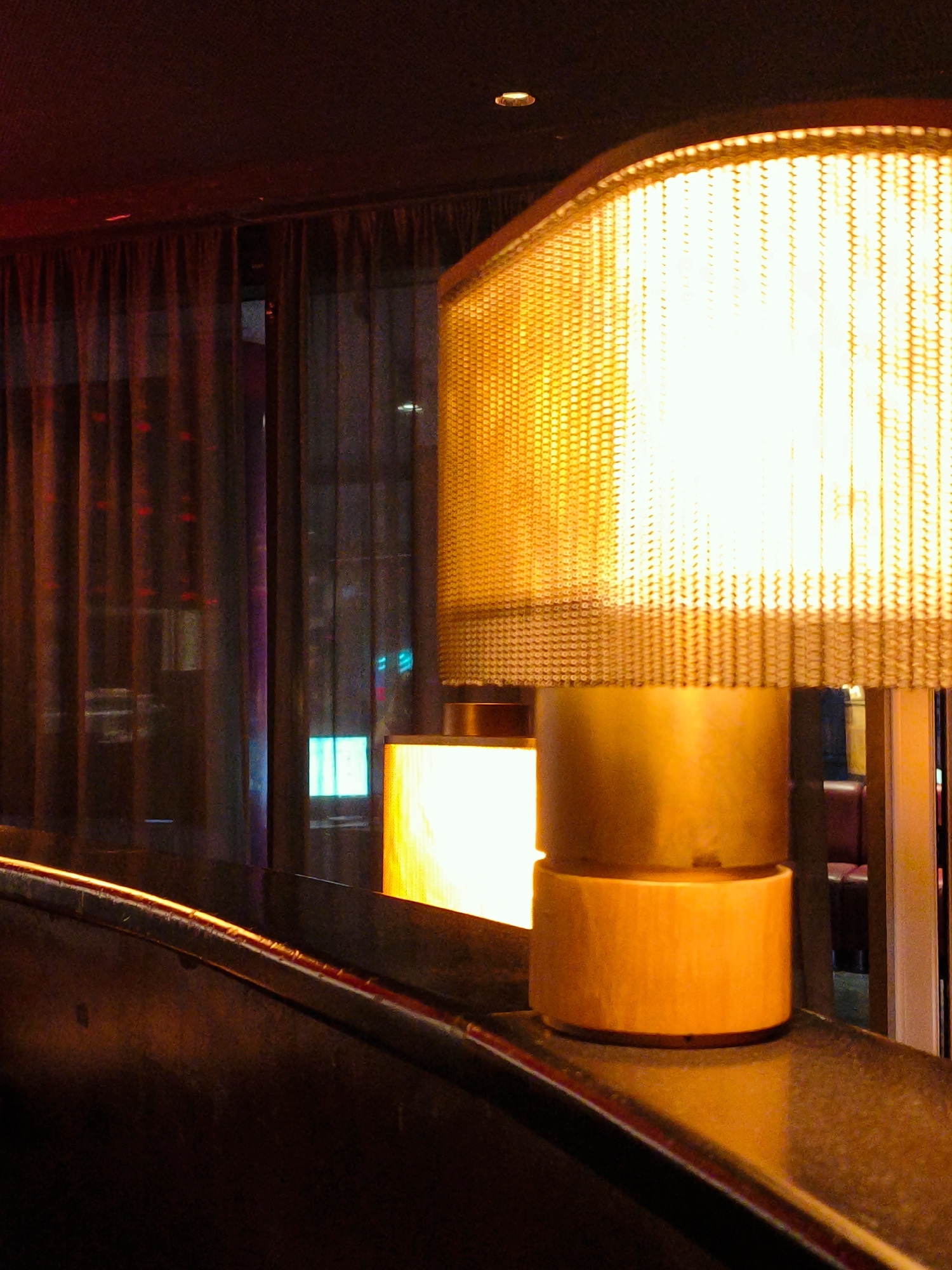 The image shows a close-up of a lamp with a golden mesh shade, emitting a bright, warm light. The lamp is positioned on what appears to be a dark, curved surface, possibly a table or a bar. Behind the lamp, dark curtains hang, obscuring the background. Through gaps in the curtains, faint reflections of city lights can be seen, suggesting an urban environment. The overall ambiance is dim and atmospheric, with the lamp being the primary source of illumination. There are no people or discernible actions depicted in the image. The image does not provide specific details to identify its location within Be'er Sheva, Israel.