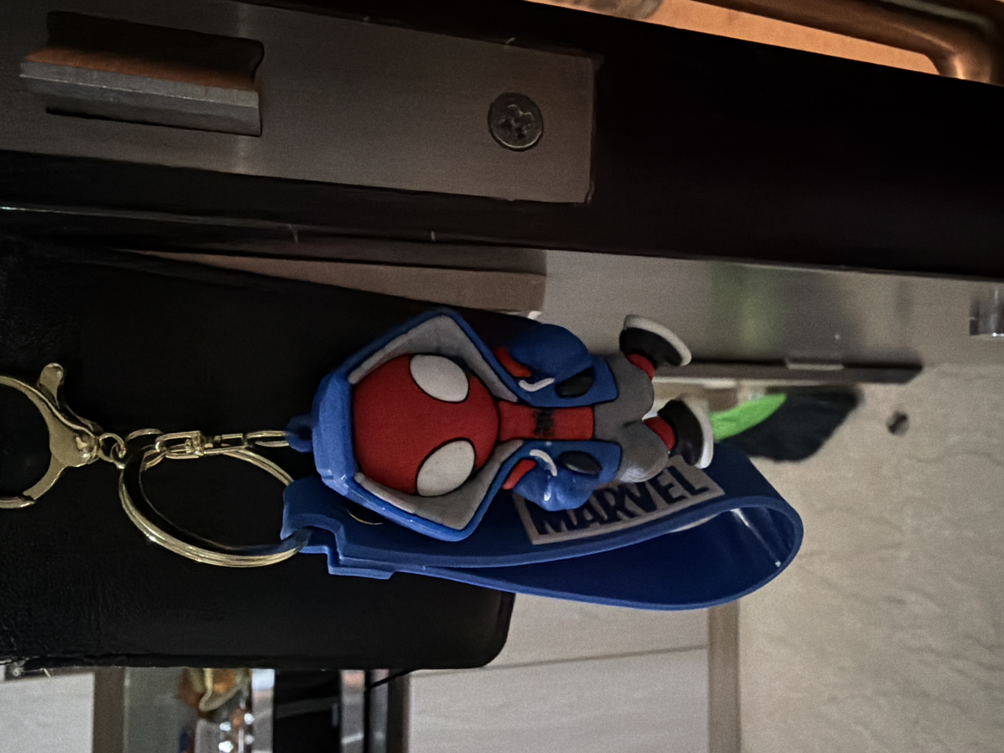 A Spiderman keychain with a blue hooded jacket and the "MARVEL" logo hangs from a metal clip attached to a black wallet. The keychain is suspended from what appears to be a door jamb in what looks like an indoor setting. The background is blurred, showing a tiled floor and a sliver of what could be a kitchen appliance. The location is Central Jakarta, Indonesia.