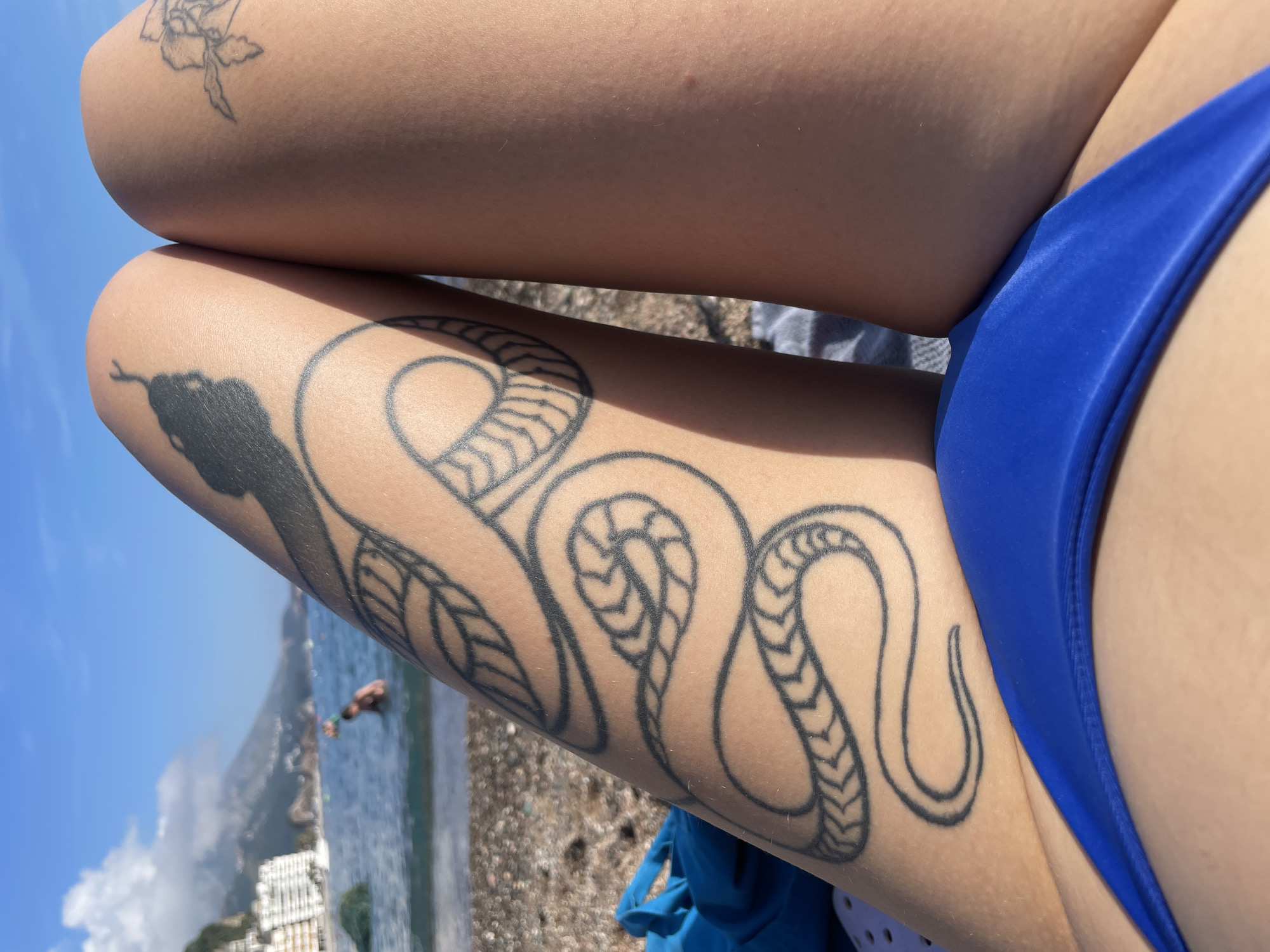The image presents a close-up view of a person's legs, featuring prominent tattoos, set against a sun-drenched coastal backdrop. The person is wearing a vibrant royal blue bikini bottom, visible along the inner thigh and hip area in the lower right.
A large, black outline snake tattoo extends across the lower leg, curving along the calf and shin. The snake's head, facing left, is solid black, while its body is characterized by detailed patterns resembling braided rope or scales. On the upper thigh, in the top right quadrant of the image, a lighter black outline floral tattoo, possibly a rose, is visible.
To the left of the legs, a scenic view of a sunny beach and the sea unfolds. The calm blue water stretches out, meeting a light-colored pebbly or sandy shore. A distant figure is seen in the water, possibly swimming or wading. Along the coastline in the background, several white buildings or structures indicate a populated area, consistent with a resort town like Rafailovići, Montenegro. The sky above is a clear, bright blue with a few scattered white clouds.
The strong lighting suggests it is daytime with bright sunshine, creating highlights on the skin and a crisp definition of the tattoos. The overall scene conveys an atmosphere of relaxation and leisure at a seaside location. No visible text is present in the image.