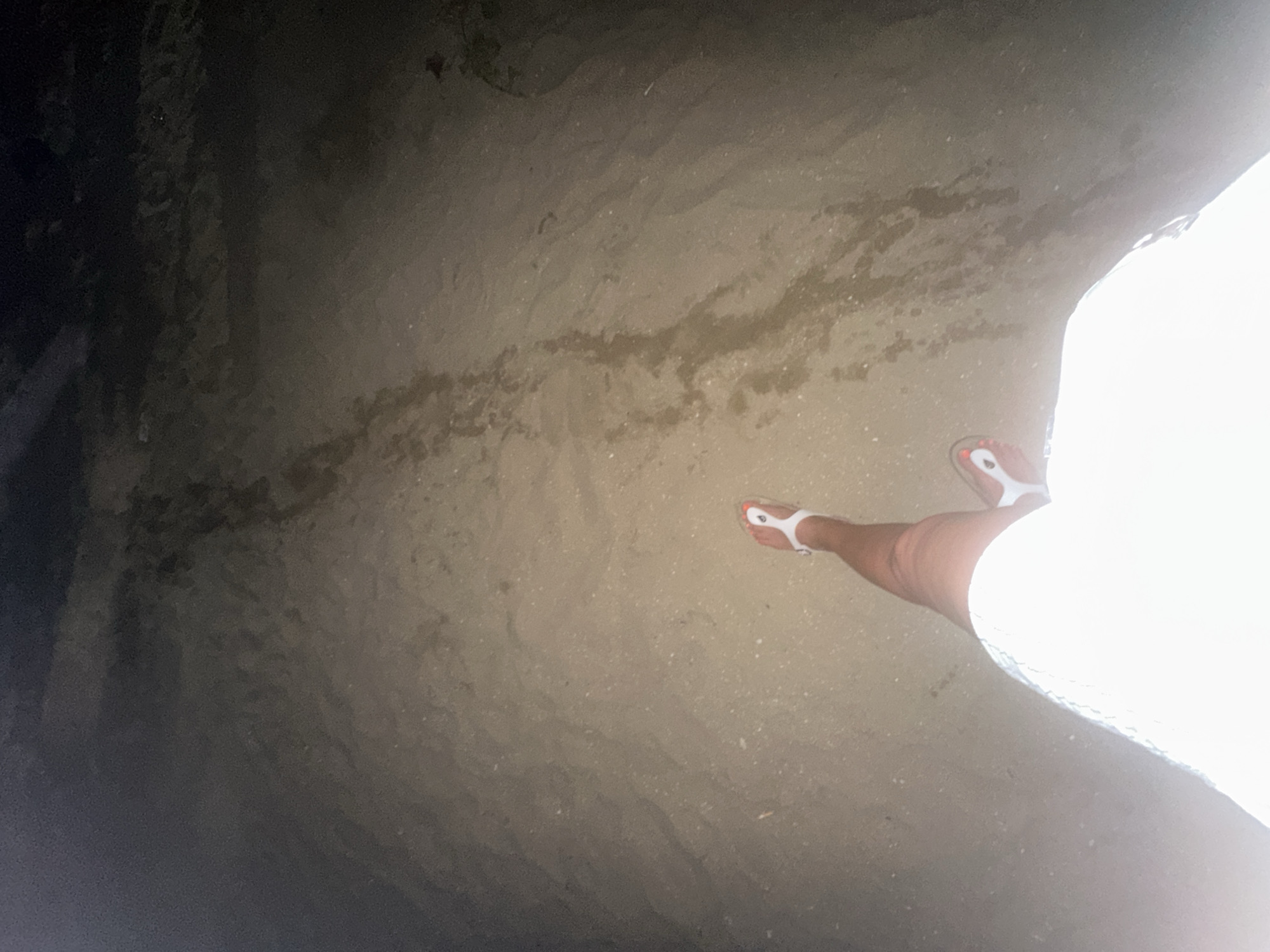 The image captures a ground-level view of a person standing on what appears to be a dark, damp sandy or muddy surface, consistent with a beach or shoreline at night in Pattaya City, Thailand. Only the lower right leg and foot of the person are visible on the right side of the frame. They are wearing white flip-flops, and their toes are adorned with bright orange nail polish. The white garment worn by the person, possibly a dress or skirt, is heavily overexposed, creating a stark contrast with the darker surroundings.
The ground exhibits irregular patterns and textures, suggesting it is wet or covered in shallow water, reflecting the low ambient light. The left side of the image is dominated by a very dark, indistinct area, likely the sea or a shaded part of the land under minimal illumination. There are no other discernible objects, structures, or visible text in the scene. The overall impression is one of a nighttime stroll along a quiet, dimly lit shore.