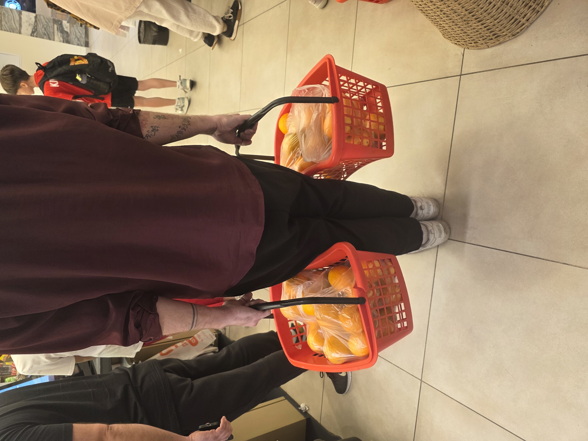 A person in black pants and a burgundy shirt carries two orange plastic baskets filled with oranges in plastic bags. The setting appears to be a supermarket or grocery store, given the tiled floor and the presence of other shoppers in the background. The location is not discernible as being in Krasnodar, Russia, from the image alone.