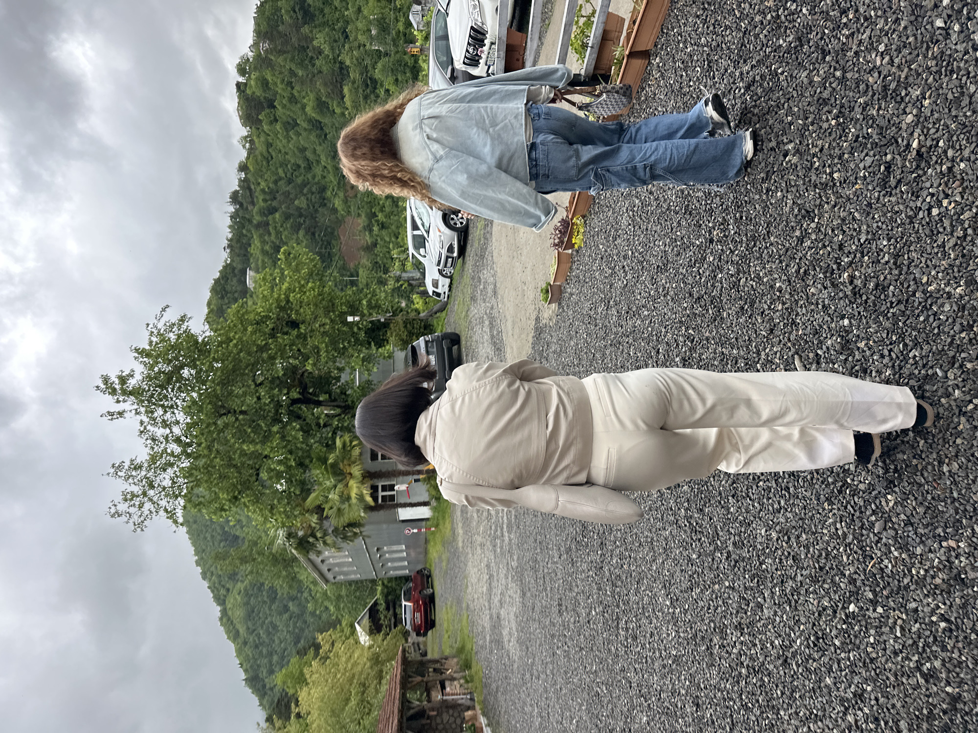 The image shows two individuals standing on a gravel surface in what appears to be a parking area. The background features a building and a heavily vegetated mountain slope. The sky is overcast.
One individual, closer to the foreground, is wearing a light-colored jumpsuit. The other, further away, is wearing a denim jacket and jeans. The two individuals are standing apart from each other.
Several cars are parked in the background. There is vegetation visible in the foreground and background. The overall environment suggests a rural or semi-rural location. Based on the location, this is likely Tibeta, Georgia.