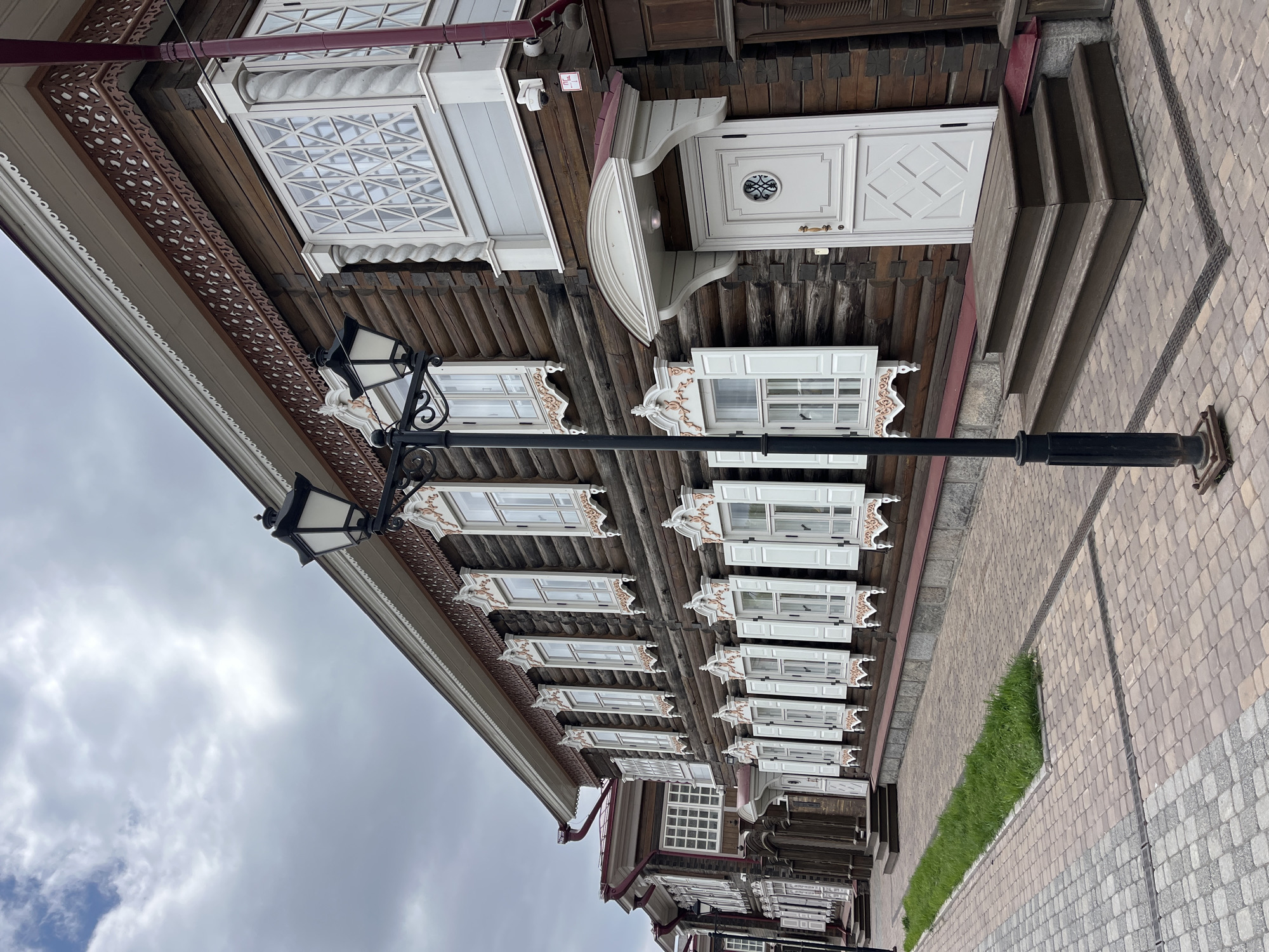 The image shows the architecture of a traditional Russian building. The building is characterized by wooden construction with elaborate decorative trim, particularly around the windows and roofline. The windows are white with detailed, ornate carvings. A black lamp post with multiple lamps stands in front of the building.
The building's exterior is made of brown wood, and there is a white door with a circular window detail above a set of stairs. The area in front of the building is paved with bricks, and there is a patch of grass growing along the edge of the pavement. The sky above is overcast with gray clouds.
Based on the architectural style and the information given, this is likely a historical building located in the city of Krasnoyarsk, Russia.