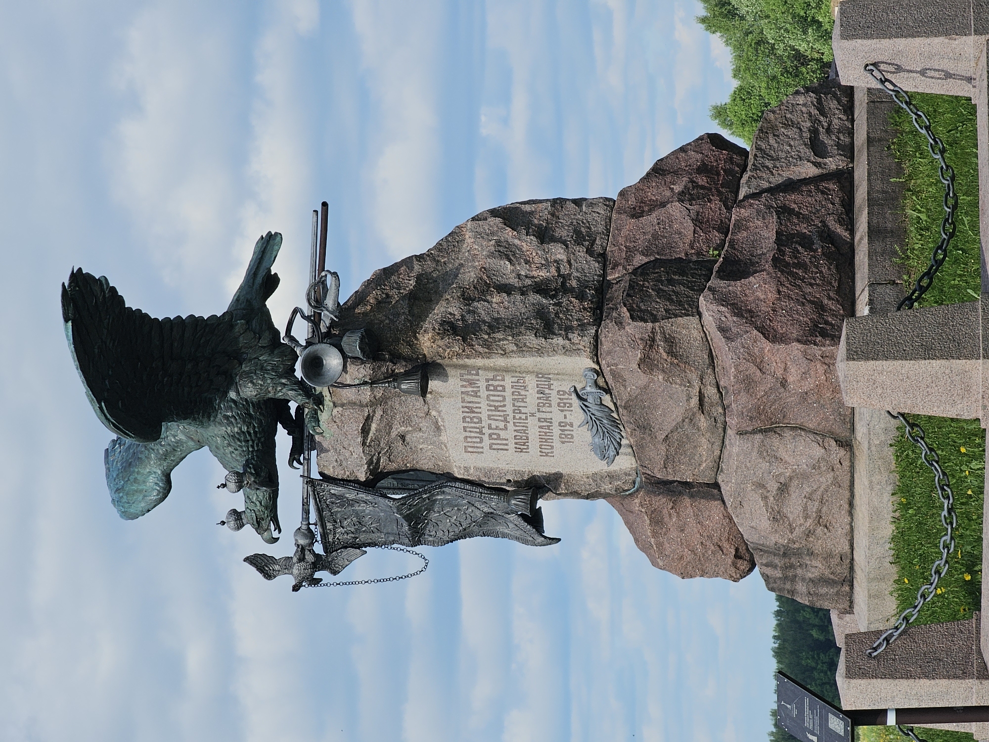 The image depicts the monument to the Cavalry Guard in Borodino, Russia, set against a backdrop of a partly cloudy sky and some greenery. The monument features a bronze double-headed eagle, topped with crowns, perched atop a rock structure. An engraved plaque is affixed to the rock, and a metal flag and other symbolic weaponry are positioned on the monument. A chain extends from a stone structure to the right of the rock formation, and foliage is visible in the background.