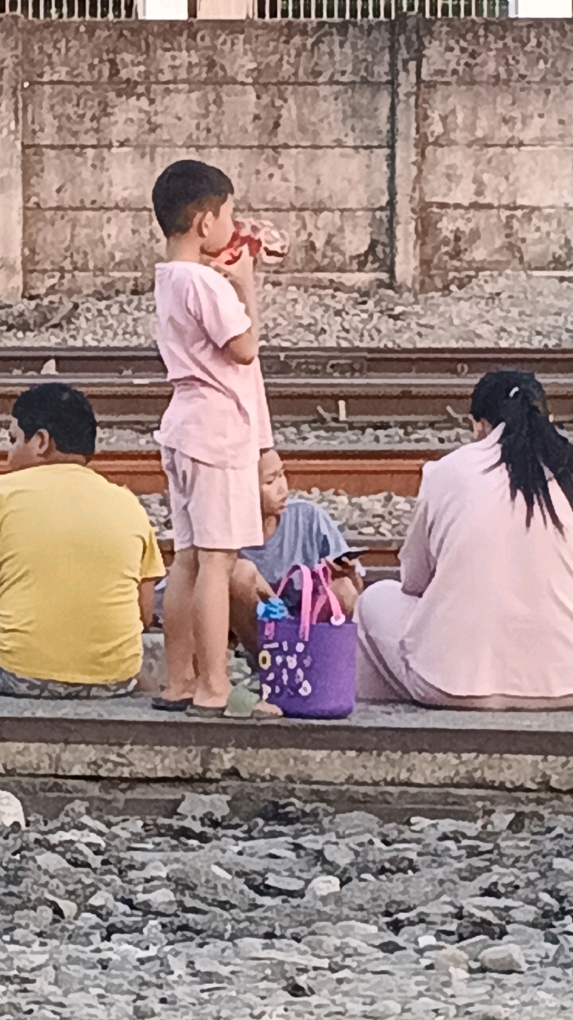 The image shows a group of people, including children, sitting by a train track. The background appears to be a concrete wall with horizontal lines. The people are casually dressed, with some wearing pink and yellow clothing. One child is drinking from a bottle. A purple bag with decorations is visible on the ground. The scene is set on what appears to be a train platform or trackside in Semarang, Indonesia. The ground is covered with gravel.